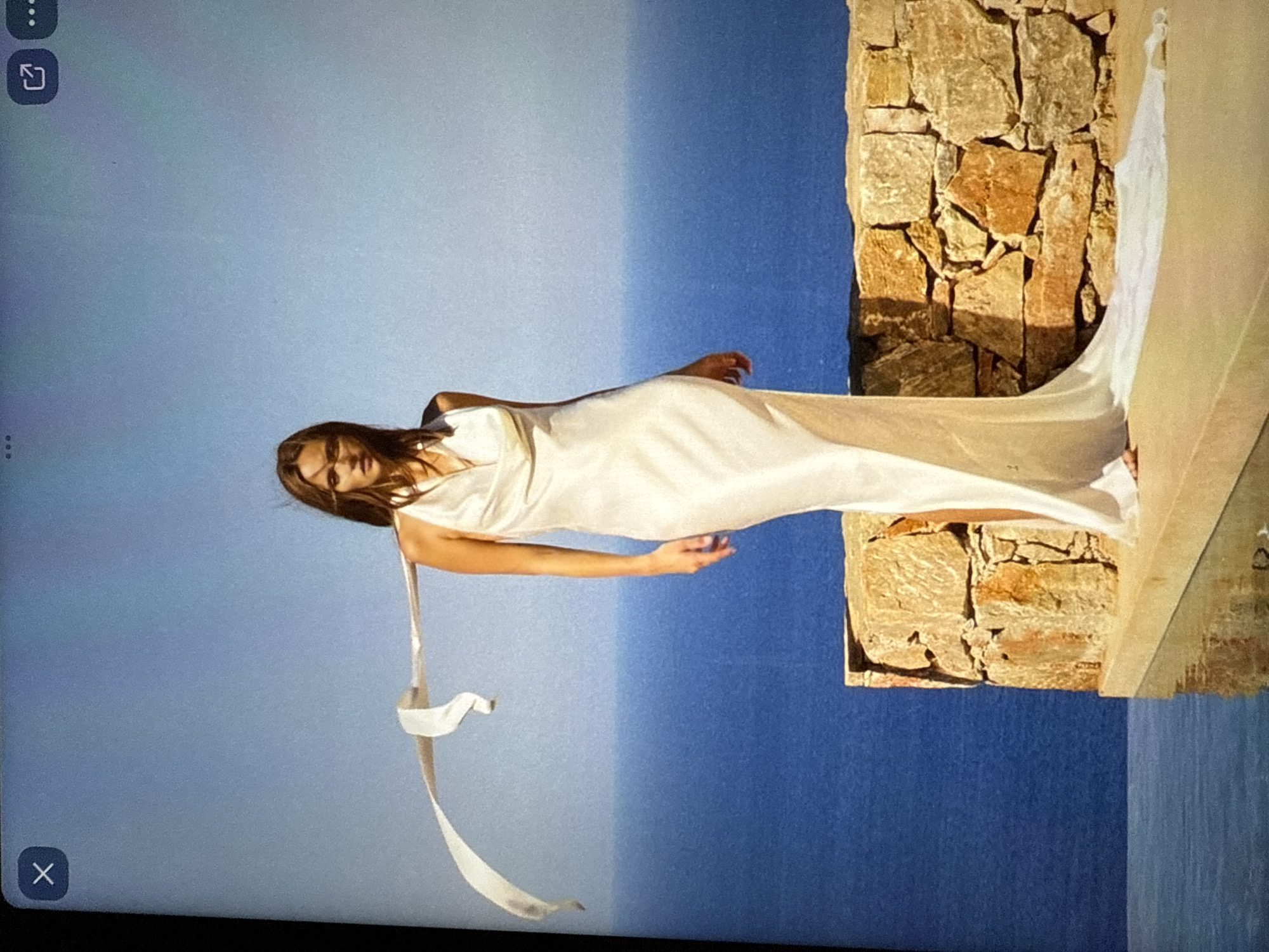 A woman stands on a stone ledge by the sea in Găvănoasa, Moldova, wearing a flowing white halter dress. A white sash trails behind her, caught in the wind. The sea is a deep blue, and the sky is a pale blue. The stone wall behind her is made of rough, irregular stones.