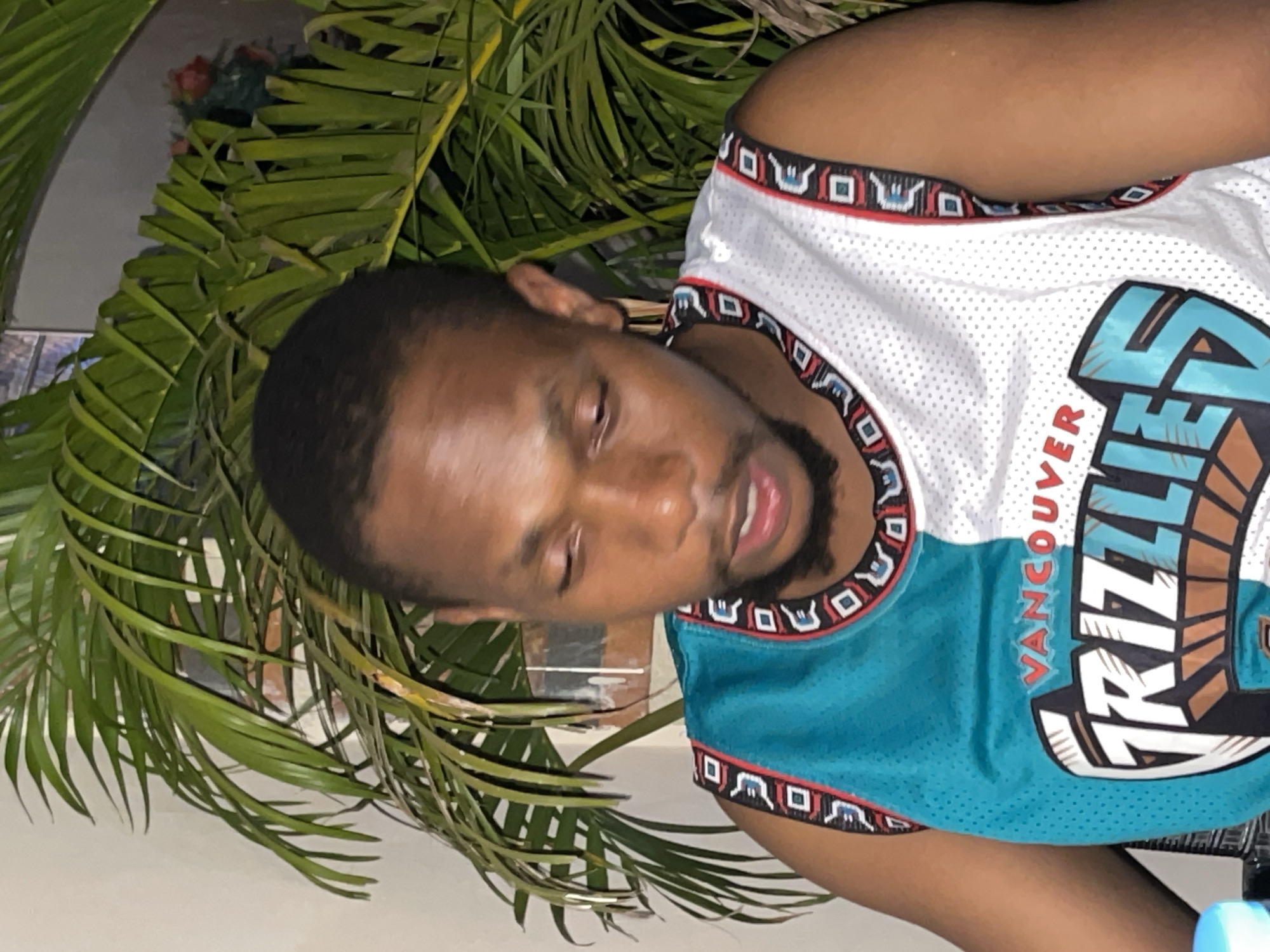 A man is wearing a Vancouver Grizzlies jersey. He is outdoors, with palm fronds in the background. There is no information to identify the city of Abuja, Nigeria in the image.