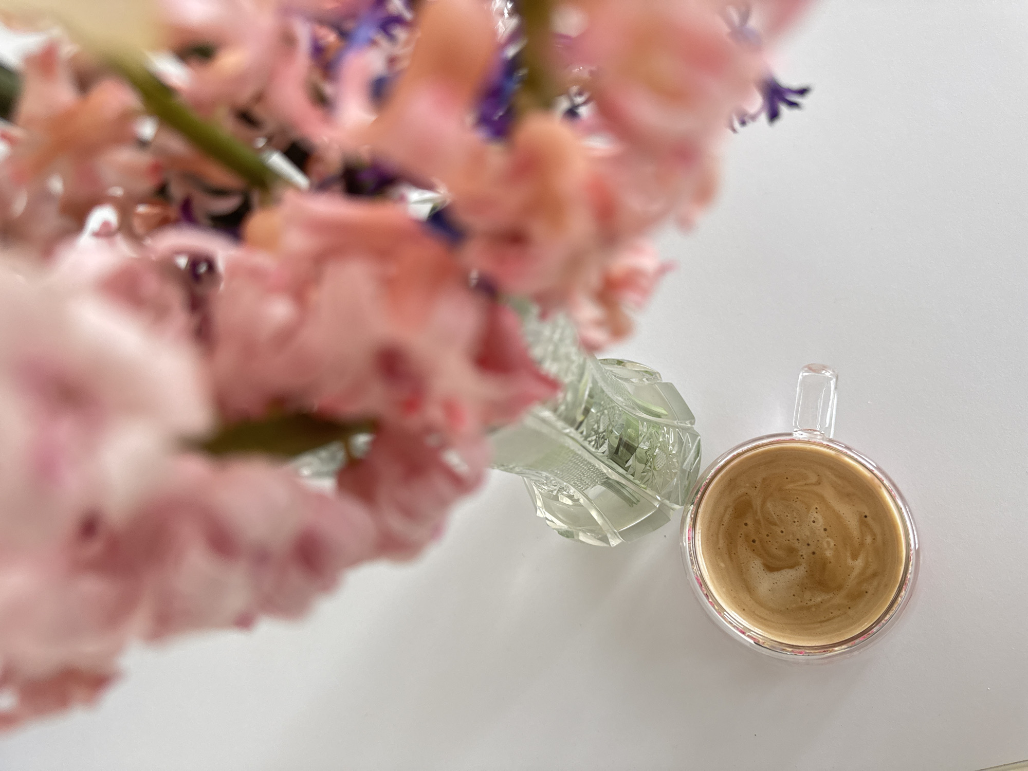 An eye-level, slightly blurred photograph shows a clear glass mug of coffee with a foamy surface next to a crystal vase containing a bouquet of pink and purple flowers. The objects sit on a white surface, possibly a table or countertop. The background is out of focus. There is no indication of the location in the city of Varna, Bulgaria. No people are visible.