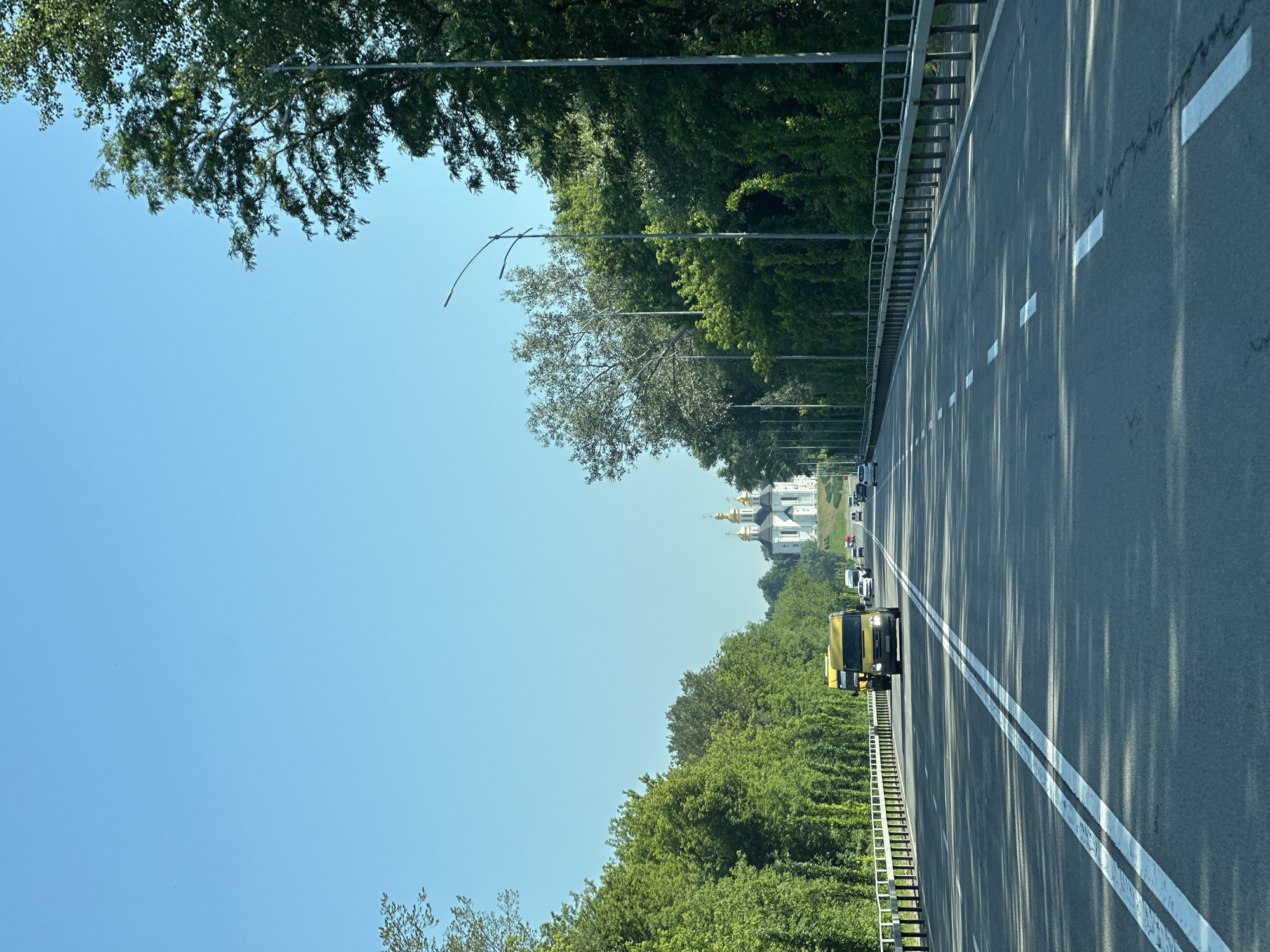 The image shows a view from a vehicle traveling on a multi-lane highway in Chernihiv, Ukraine. The highway has distinct white lane markings. On either side of the road, dense green trees line the landscape, with some taller trees extending towards the clear blue sky. A white metal guardrail runs along the edge of the road. In the distance, a white church with golden domes is visible on a hill. Several vehicles, including a yellow bus and cars, are on the highway. Streetlights are also present along the roadside. The sun is casting shadows on the asphalt, indicating it is daytime.