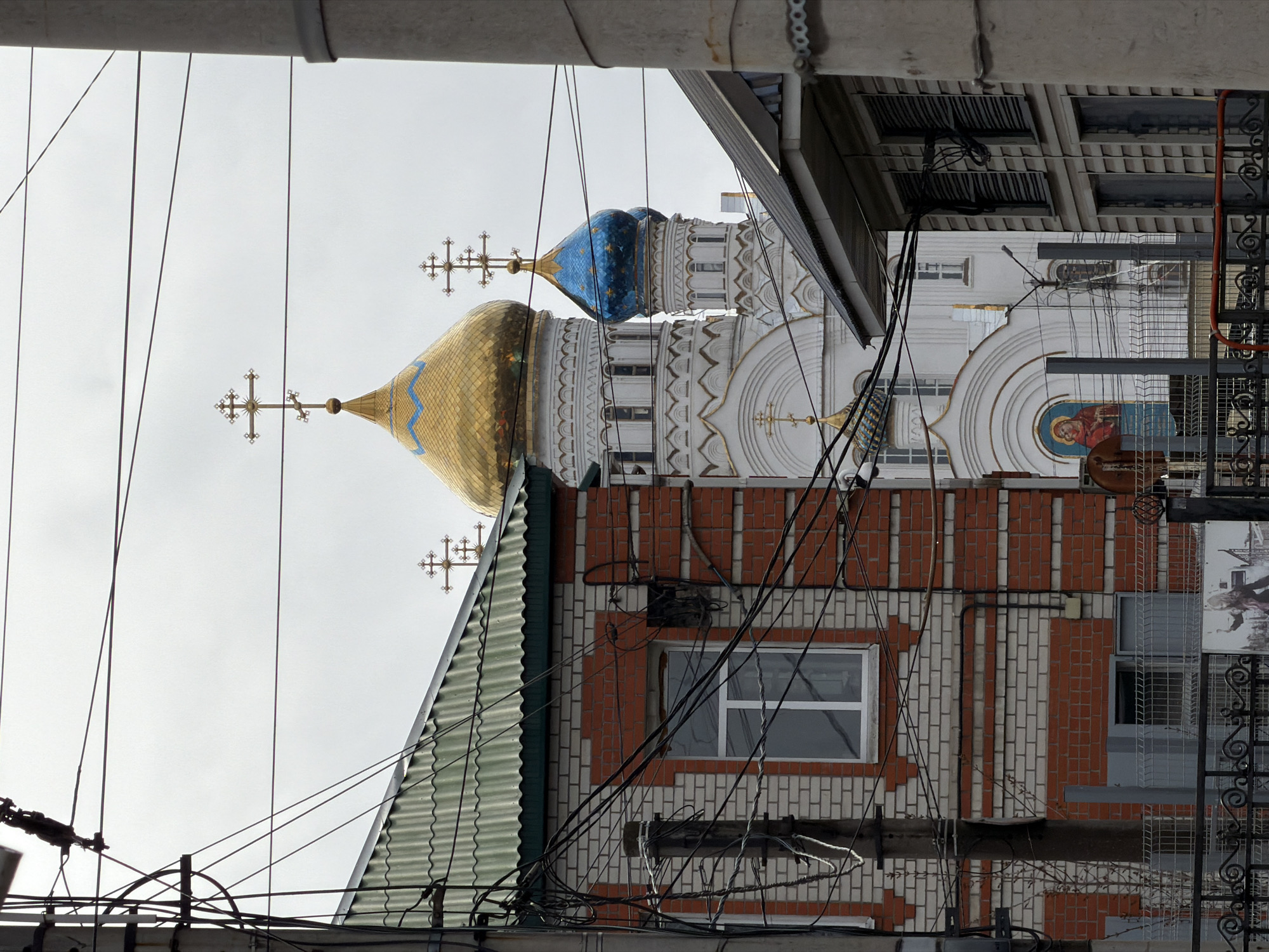 This vertically oriented image captures a dense urban scene in Liski, Russia, featuring a prominent Orthodox church and a neighboring brick building, both partially obscured by a network of overhead electrical wires.
In the background, a white Orthodox church stands out with its ornate architecture. It features a large, shimmering golden dome adorned with a blue patterned band, topped by an intricate golden Orthodox cross. To its right, a smaller, dark blue dome with stars is also visible, similarly crowned with a golden cross. The church facade displays decorative white and gold molding, arches, and a religious icon painting depicting a figure in red and blue robes.
In the mid-ground and foreground, a brick building with a distinctive pattern of alternating red and white bricks is visible. Part of its green corrugated roof is seen below the church's golden dome. A white-framed window is set into the brick wall. Numerous black electrical wires and cables crisscross the entire frame, forming a dense web, particularly concentrated in the lower and right sections of the image. These wires are connected to various fixtures and what appears to be a wooden beam. Elements of metal railings or fences are also visible on the right.
The sky is uniformly overcast and light grey, suggesting a daytime setting with soft, diffused light. There is no visible human activity or legible text in the image. The composition highlights the juxtaposition of traditional religious architecture with the pervasive infrastructure of a modern town.