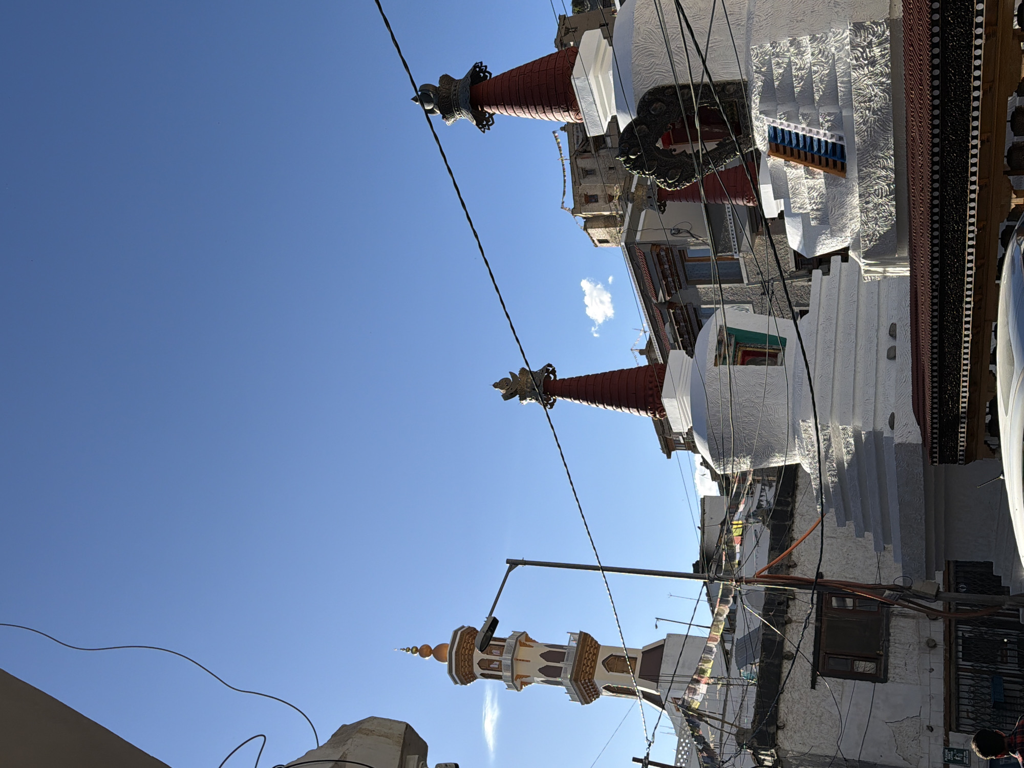 The image, taken in the city of Leh, India, features a collection of stupas and a minaret under a clear blue sky. The foreground is dominated by the white, tiered structures of several stupas, with some adorned with red conical tops and golden finials. One stupa shows a blue and yellow set of stairs leading to an opening. To the left, a tall, slender minaret with white and gold detailing stands prominently, topped with a golden spire. Prayer flags, strung between buildings, add a splash of color to the scene. Buildings with traditional architecture, constructed from stone and plaster, are visible in the background. Electrical wires crisscross the sky, and a street lamp is positioned near the minaret. The lower part of the image shows a section of a building with a window and a barred entrance, and a person partially visible wearing a red and black checkered shirt. A car's roof is also visible in the bottom right corner. The overall impression is one of a spiritual and culturally rich urban landscape.