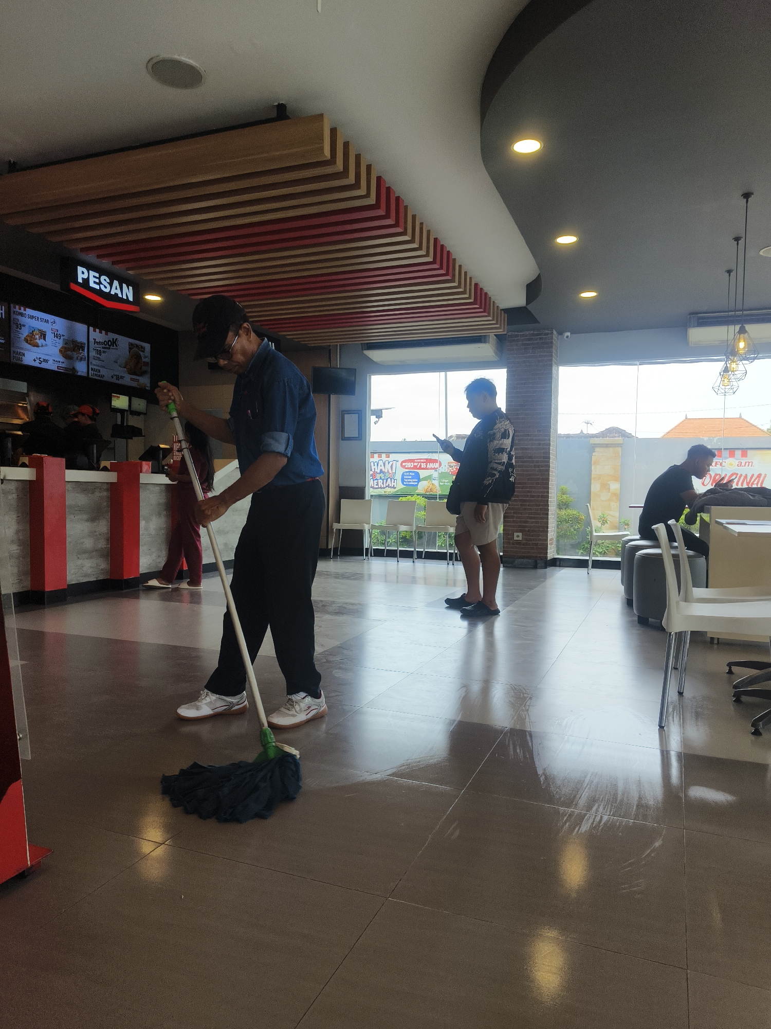 The image depicts the interior of a KFC restaurant, likely located in Denpasar, Indonesia, as suggested by the signage which appears to be in Indonesian. A male employee, wearing a dark blue shirt, dark pants, and a baseball cap, is actively mopping the tiled floor with a blue mop. He is positioned in the foreground, facing away from the camera, with his attention focused on cleaning. In the background, another person is visible at the counter, potentially a customer or another staff member. Further in the background, two other individuals are seated, one looking at their phone and the other at a table. The restaurant environment features modern lighting, a suspended ceiling with decorative wooden slats, and large windows allowing natural light to enter. The overall scene captures a moment of daily operation within a fast-food establishment.