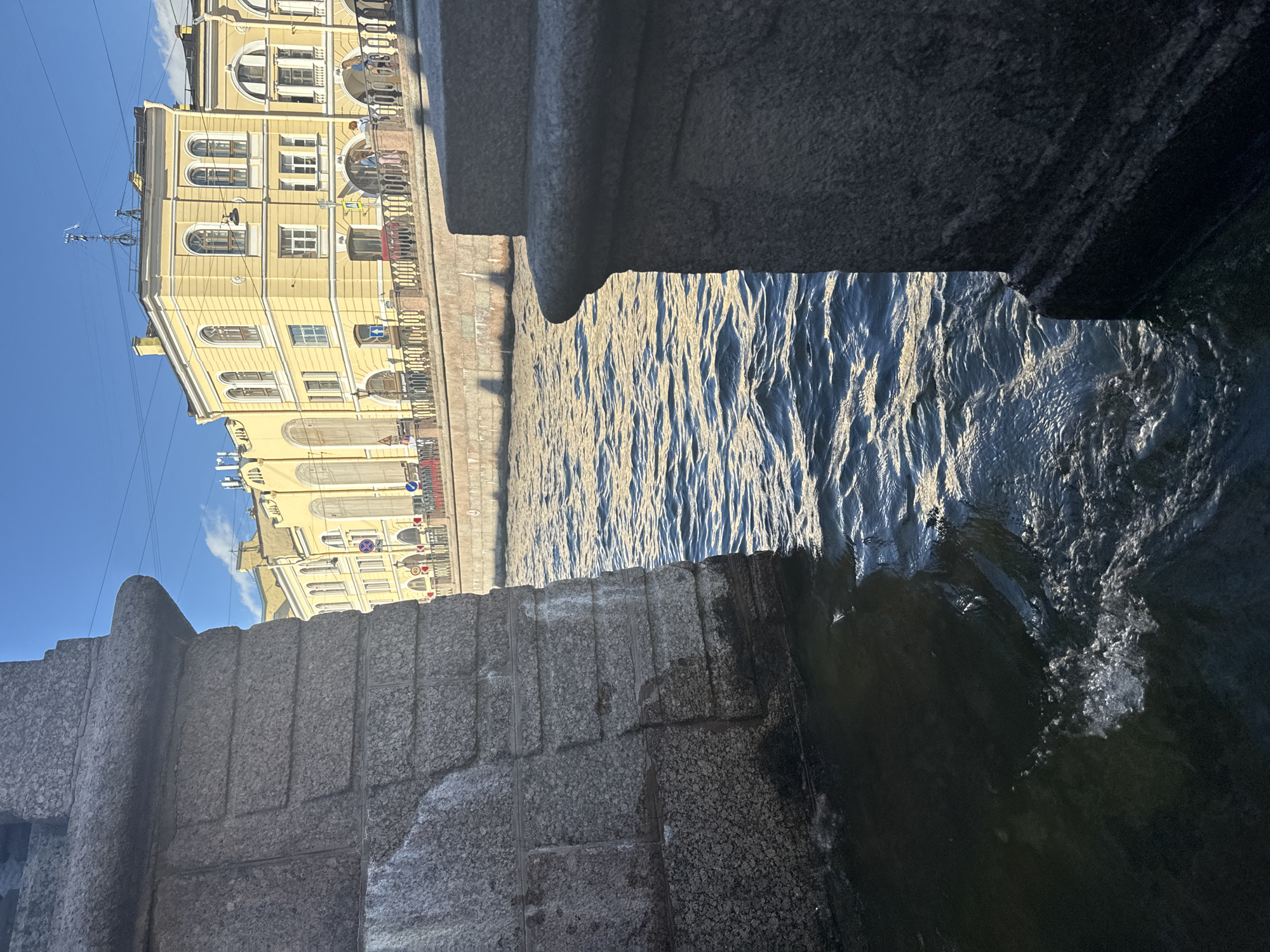The image shows a view of a canal in Saint Petersburg, Russia, with a yellow building in the background and the stone supports of a bridge in the foreground. The canal water is choppy and reflects the sunlight. A few people are visible walking along the embankment in front of the building. The building has multiple arched windows and a detailed facade, characteristic of St. Petersburg architecture. The sky is clear and blue. The stone supports in the foreground are large and rough-hewn, suggesting they are part of an old bridge.