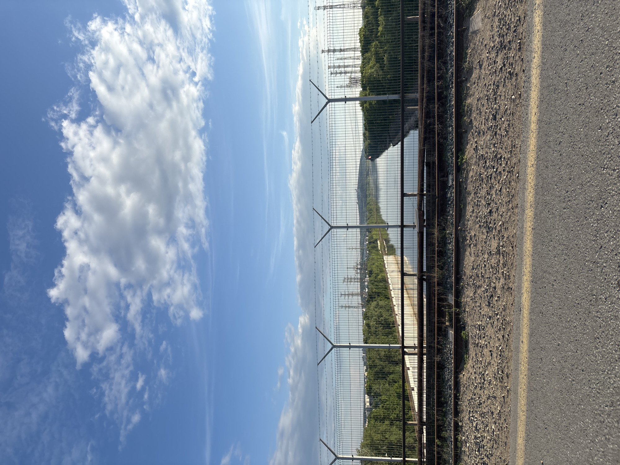 This image presents an elevated, static view over a waterway in what is identified as Chaikovsky, Russia. The scene is dominated by a clear blue sky with scattered white cumulus clouds. In the foreground, an asphalt road with a faded yellow line on its right edge leads to a section of unused railway tracks, characterized by gravel ballast and two rusty metal rails. Above the tracks, a prominent metal chain-link fence, topped with barbed wire and supported by Y-shaped posts, spans horizontally across the frame.
Beyond the fence, a calm, reflective canal or river extends into the distance, bordered on both sides by dense green trees and a visible concrete embankment on the left. On the distant horizon, urban buildings are discernible on the left, partially obscured by trees, while on the right, multiple tall industrial structures, resembling power line towers or factory chimneys, rise above the tree line. No people or active interactions are visible in the scene. The overall environment suggests an industrial or utility area adjacent to an urban settlement, with a focus on infrastructure and natural elements.