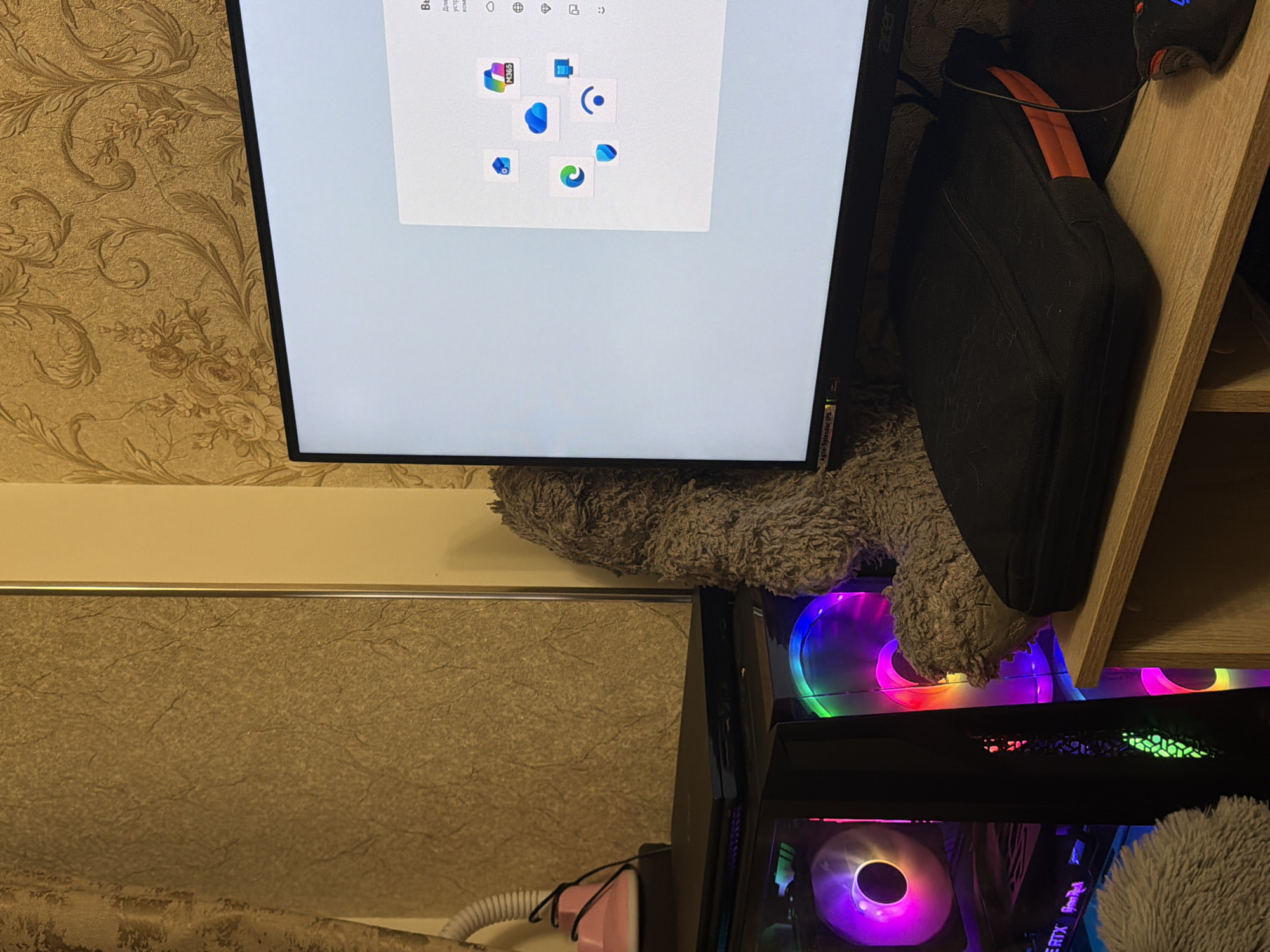 The image depicts a computer setup indoors, with a monitor displaying a Microsoft 365 prompt and an Acer logo visible in the top left corner. A black bag with an orange strap rests against a wooden shelf and sits atop a grey fluffy cushion. The computer tower next to the monitor features visible RGB lighting. The wallpaper behind the monitor has an intricate floral pattern. A portion of the scene includes a white corrugated hose and a pink object, and there's a cabinet or shelf visible underneath the desk. It is not possible to determine the location of the image as Yerevan, Armenia.