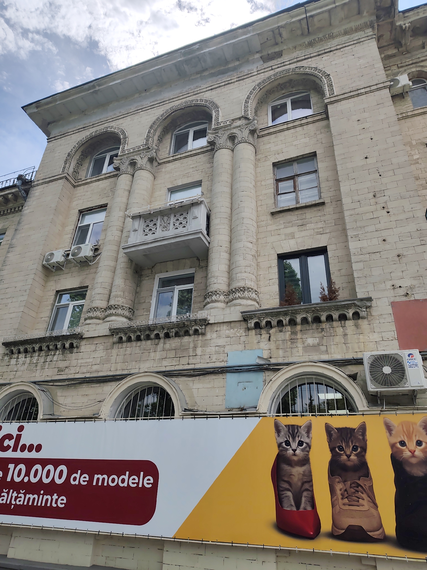 The image shows a building in Chișinău, Moldova, with a large billboard in front. The building is constructed of light-colored stone blocks and features arched windows, columns, and decorative elements. Several air conditioning units are visible on the facade. The billboard is divided into two sections: a white and red section with text that appears to be an advertisement, and a yellow section featuring an image of three kittens. The kitten on the left is inside a red high-heeled shoe, the middle kitten is inside a brown sneaker, and the kitten on the right is wearing a black garment. The sky above is cloudy. The billboard is attached to the lower part of the building.