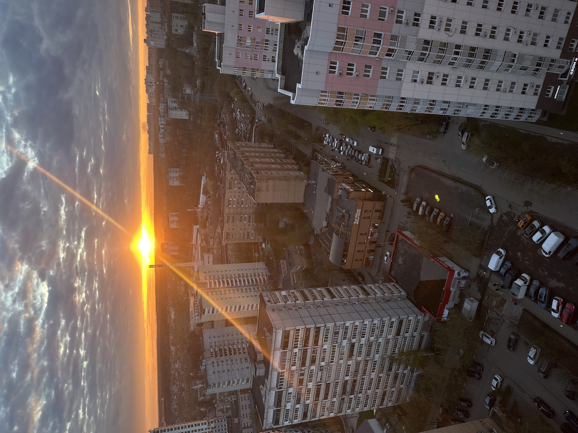 The image depicts a high-angle, oblique view of a cityscape at sunrise. A bright sun is visible, emanating strong rays of light. To the right of the sun, dense clouds are visible, and to the right of the clouds is a dense forest. There are many cars lining the streets. There are also buildings, all of which seem to be high-rise. There is a sign that reads "OZON." Based on the street layout and signage, the location is likely the city of Khimki, Russia.
