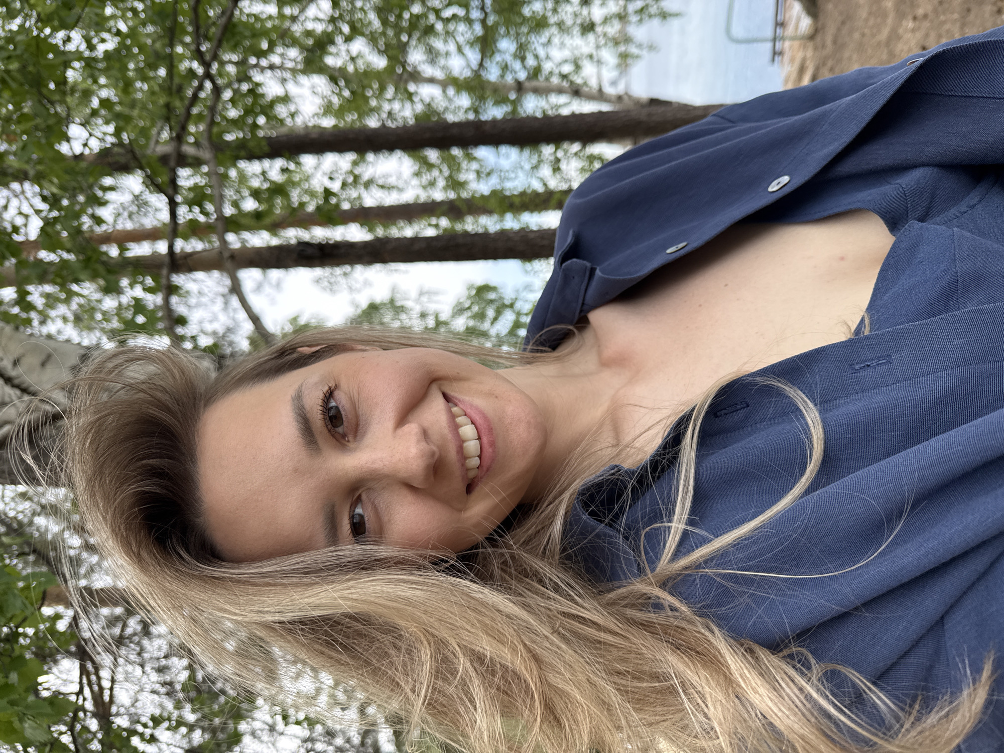A woman with blonde hair and fair skin smiles at the camera. She is wearing a dark blue button-down shirt. The background includes trees and a body of water, suggesting an outdoor location. The setting may be near the city of Chaikovsky, Russia, though this is difficult to confirm without more information.