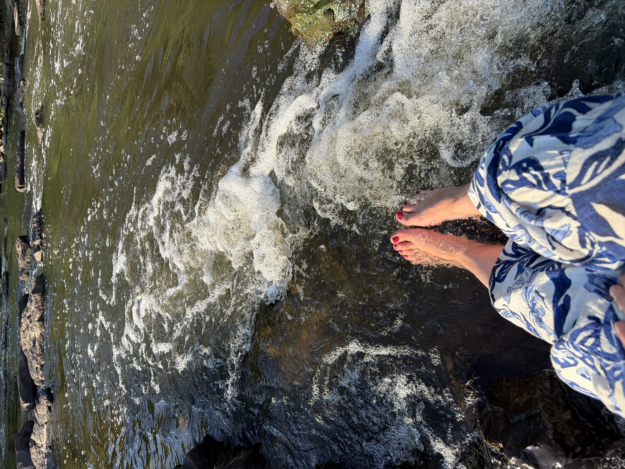 This image captures a person's bare feet, painted with red nail polish, dangling in the flowing water of the Glan river in Meisenheim, Germany. The water is a murky green, with white foam and bubbles indicating a gentle current and ripples. Rocks of various sizes are scattered in the river, with some visible on the banks. The person is wearing patterned pants with blue and white floral designs. The sunlight reflects off the water, creating a sparkling effect. The overall scene suggests a moment of relaxation and connection with nature in a natural outdoor setting.