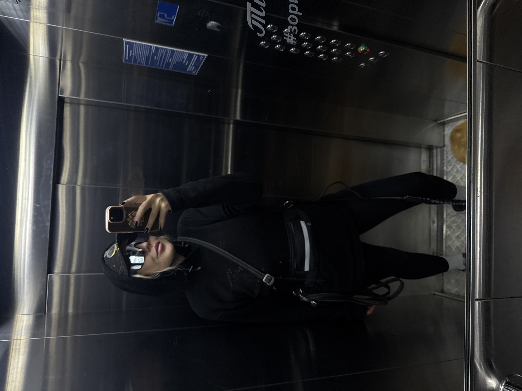 A person in dark clothing takes a mirror selfie inside an elevator. The elevator has a panel with buttons and writing in Cyrillic. The reflection shows the elevator's interior and door. The photo was taken in Krasnoyarsk, Russia.