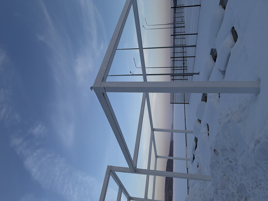 The image depicts a snow-covered waterfront scene in Chaikovsky, Russia, likely along the Kama Reservoir. In the foreground, multiple white, square-profile metal structural frames, resembling pergolas or an unfinished canopy, are partially buried in deep snow. One frame in the mid-right features a small, dark light fixture mounted on its top beam. Irregular mounds of snow cover what appear to be benches or concrete blocks beneath these structures. Beyond the frames, a dark metal fence with wire mesh panels runs horizontally across the midground. Behind the fence, several tall, grey streetlights with angled luminaires stand upright. The background features a vast, flat expanse of a frozen, snow-covered body of water, with a dark tree-lined horizon visible in the distance. The sky above is clear, transitioning from a light blue overhead to a pale pink and orange hue near the horizon, suggesting dawn or dusk. No people, subjects, or active interactions are present in the scene.
