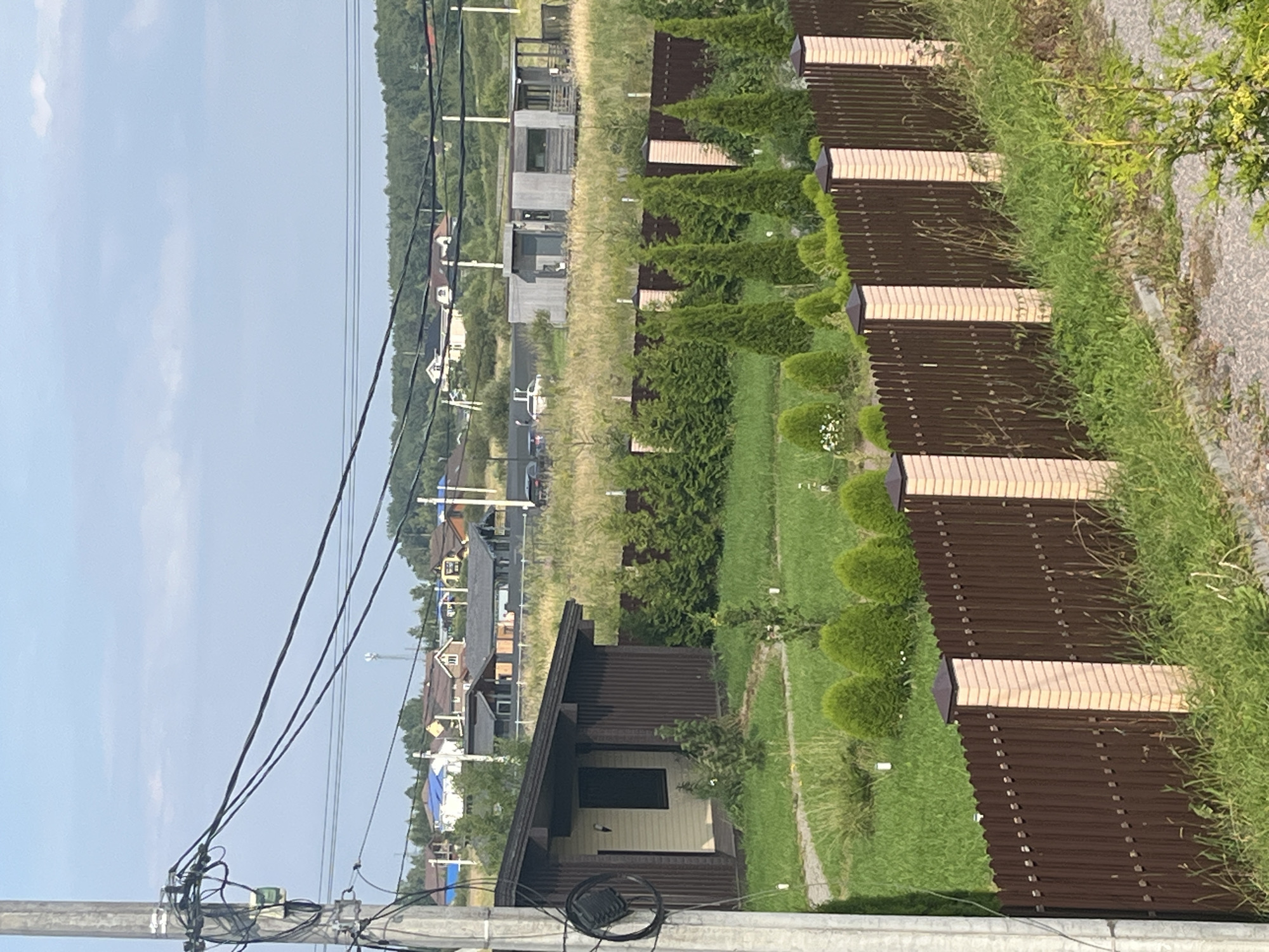 The image depicts a suburban or rural landscape in Vladimirovka, Russia, on a clear day. In the foreground, a brown wooden fence with light-colored brick posts runs horizontally across the frame. Behind the fence, a well-maintained lawn is visible, dotted with small, rounded evergreen bushes and a few young trees. A small, single-story house with a dark brown corrugated metal siding and a lighter beige section is situated to the left of the frame, partially obscured by a utility pole and electrical wires that dominate the left side of the image.
Further into the background, beyond the fence and the first house, is a field of tall, dry grass. Several more houses, of varying architectural styles and colors, are scattered across the landscape, some with dark roofs and light-colored walls. A modern-looking concrete building with large windows and a wooden deck is also present in the middle ground. In the far background, a dense line of trees forms the horizon, under a pale blue sky with scattered clouds. The overall scene suggests a residential area undergoing development or a newly established community. There are no people visible in the image.