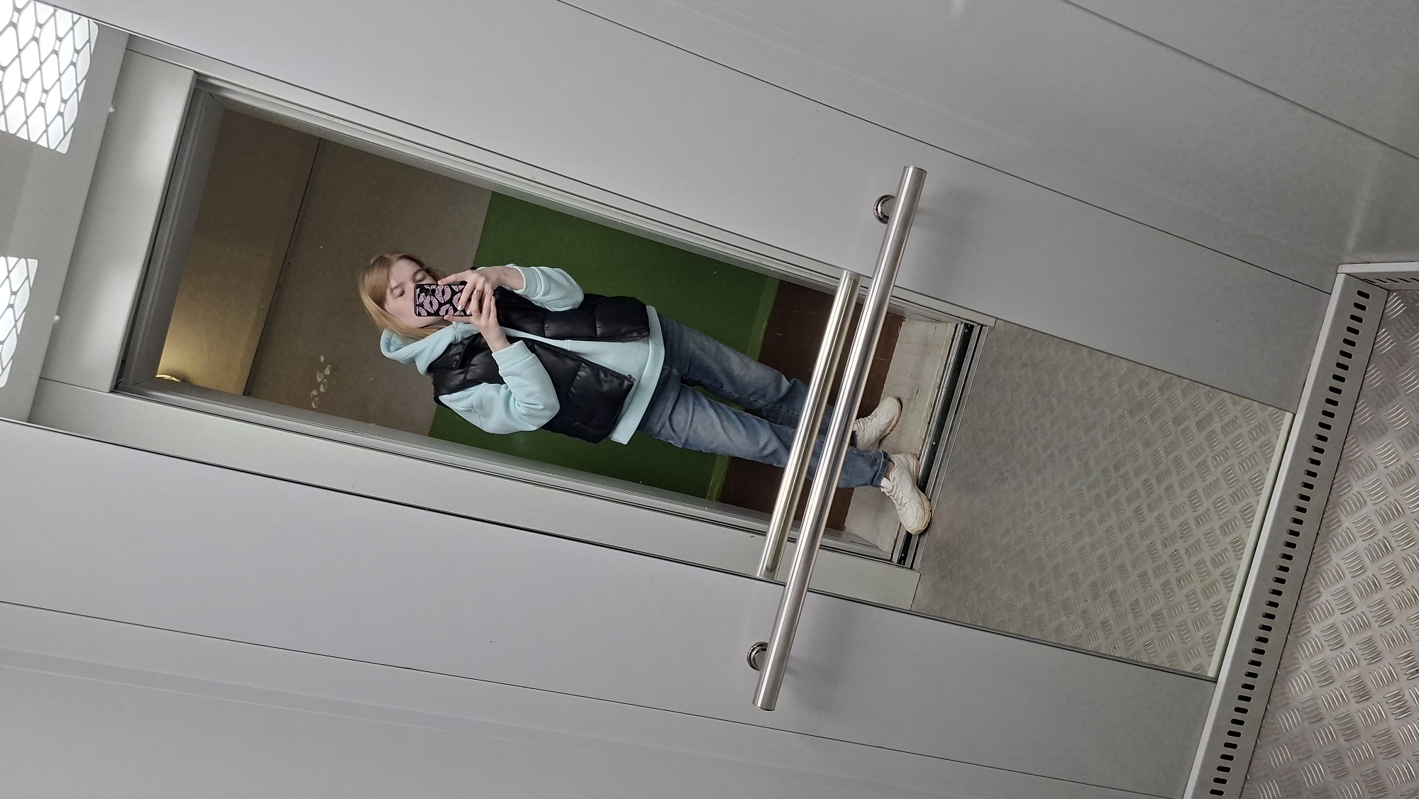 A woman is taking a mirror selfie inside an elevator or lift. She is wearing a light blue hoodie, a black vest, blue jeans, and white sneakers. Her phone case is decorated with pink lips. The interior of the elevator or lift has gray walls and a metal floor with a diamond pattern. Outside the elevator, a room with a green floor can be seen. Based on metadata, this image was taken in the city of Chaikovsky, Russia. The selfie is taken at an angle, and the details of the surrounding location are limited to what can be seen through the elevator opening. The stainless steel elevator handrails are vertically installed.