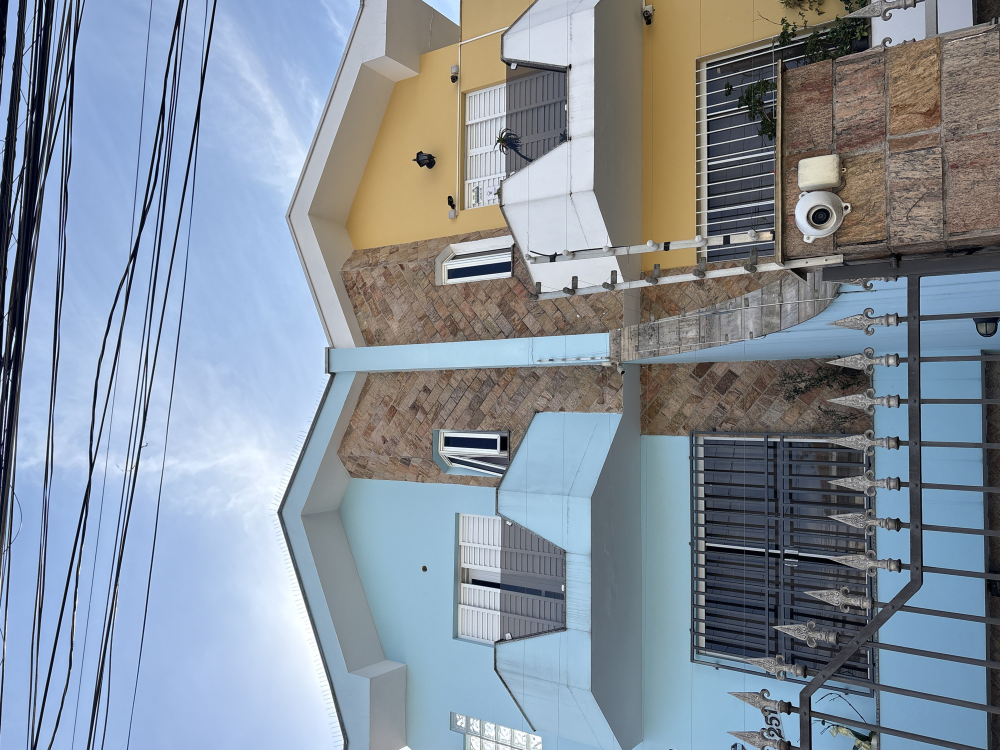 This is a low-angle, exterior, eye-level shot of two attached multi-story houses, one painted light blue and the other bright yellow, situated in the city of São Paulo, Brazil. The houses share a central vertical facade adorned with a mosaic of stone tiles. Both structures have white, sloped roofs, adorned with bird spikes. The blue house features a balcony with a pale blue railing and large, barred windows. The yellow house also has a balcony with a white railing and barred windows. Multiple black electrical wires are strung across the top of the frame against a partially cloudy blue sky. Security features are visible, including a surveillance camera mounted on a stone-clad pillar on the right side of the frame, and what appears to be an electric fence installed along the top of a fence or wall in the foreground. Decorative metal fencing with pointed finials is also present in the lower portion of the image. The overall impression is of a residential street in an urban environment.