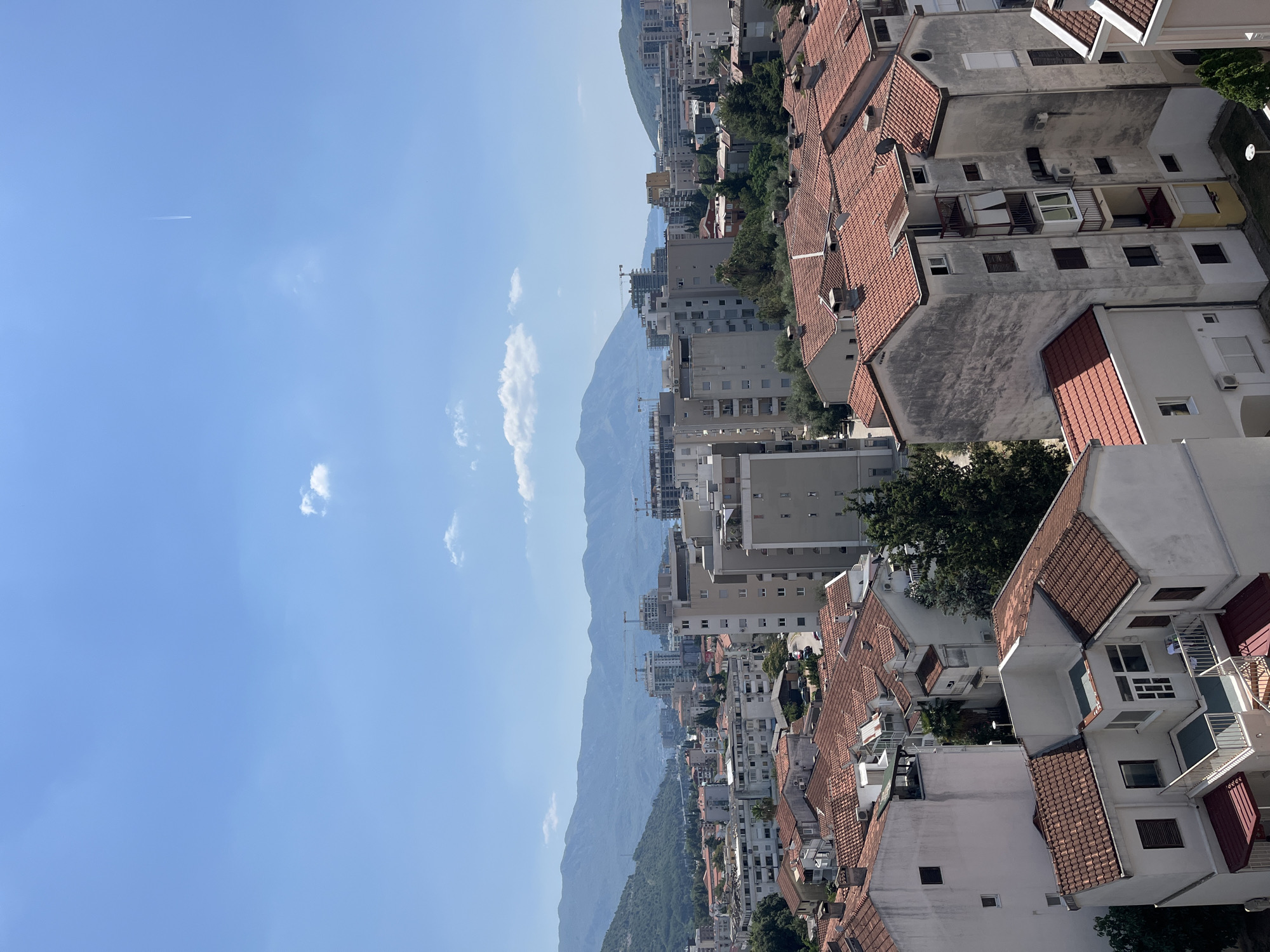 This is a high-angle, eye-level shot of a cityscape in Budva, Montenegro, on a clear, sunny day. The foreground is dominated by a dense cluster of residential buildings with red-tiled roofs. These buildings vary in height and architectural style, with some appearing older and weathered, while others are more modern. Many of these structures feature balconies and visible air conditioning units.
In the mid-ground, more buildings of varying heights are visible, interspersed with trees and greenery. The urban landscape extends into the distance, where several taller, modern buildings are under construction, identifiable by cranes. Beyond the city, a range of mountains forms a dramatic backdrop, their slopes covered in vegetation. The sky above is a vibrant blue with a few scattered white clouds and a faint airplane contrail. The scene captures a snapshot of urban development within a mountainous coastal environment.