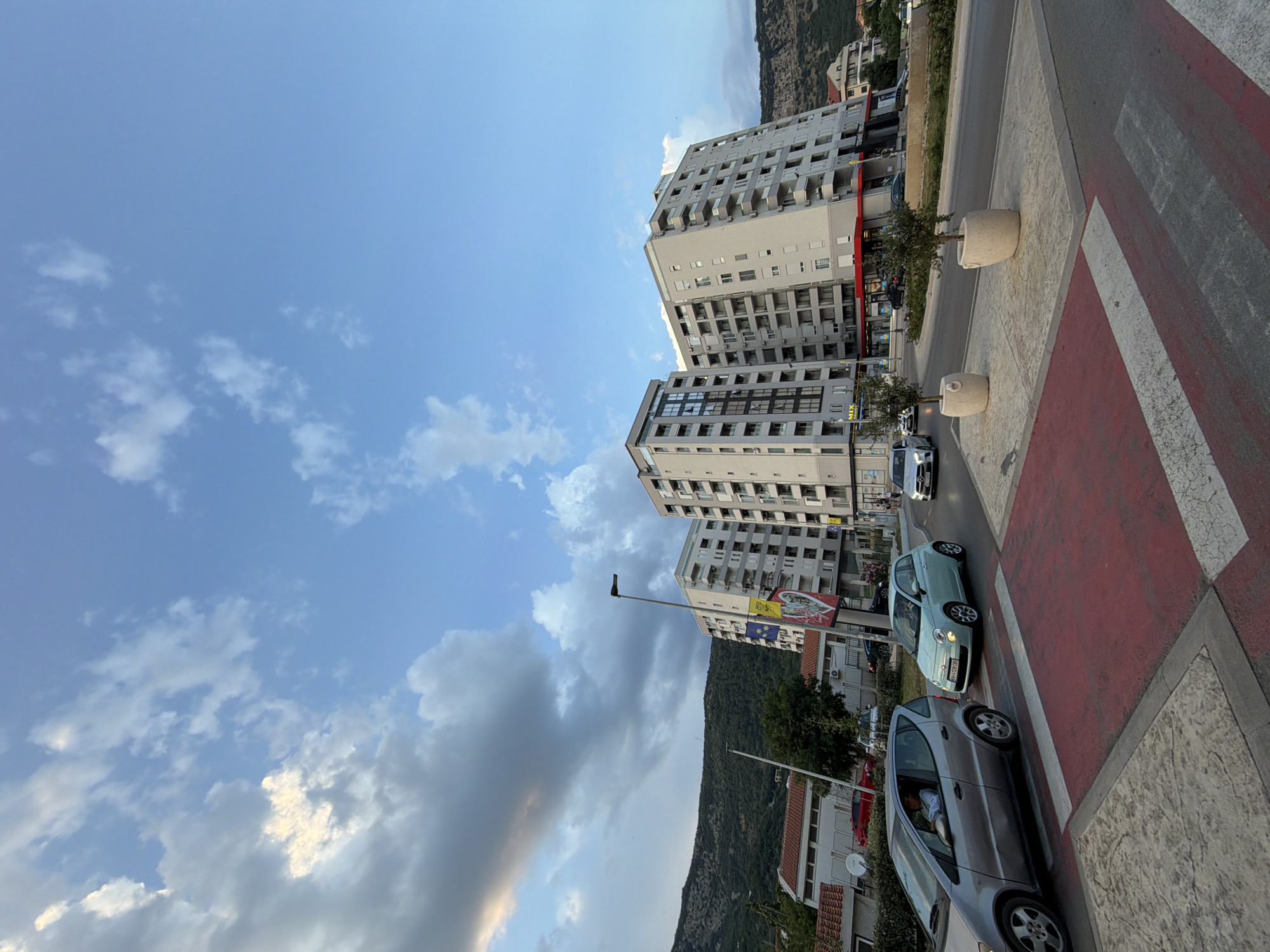 The image captures a street scene in Budva, Montenegro, under a bright, partly cloudy sky, suggesting it's daytime, likely late afternoon or early evening due to the warm light.
The primary subjects are several modern, multi-story apartment buildings lining a street. The buildings are contemporary in design, with numerous balconies and windows. A pedestrian crossing with red and white stripes is prominent in the foreground, indicating a public urban space.
Several cars are visible on the road, including a silver hatchback and a light blue compact car, suggesting normal urban traffic. One of the cars has a driver visible inside. Two large, cylindrical planters with small trees are placed along the sidewalk.
In the background, a hilly, vegetated mountainside rises, providing a natural backdrop to the urban development. The sky is a clear blue with scattered white clouds, some of which appear to be catching the sunlight, creating a sense of depth.
Text is visible on some of the buildings, including advertisements or signage, and on the license plates of the cars. One building has flags displayed, including what appears to be the European Union flag. Another sign shows a depiction of a city and flags.
The overall impression is a sunny day in a coastal Montenegrin town, with a blend of modern architecture and natural landscape. There are no overt emotional cues, but the scene conveys a sense of everyday life and activity.