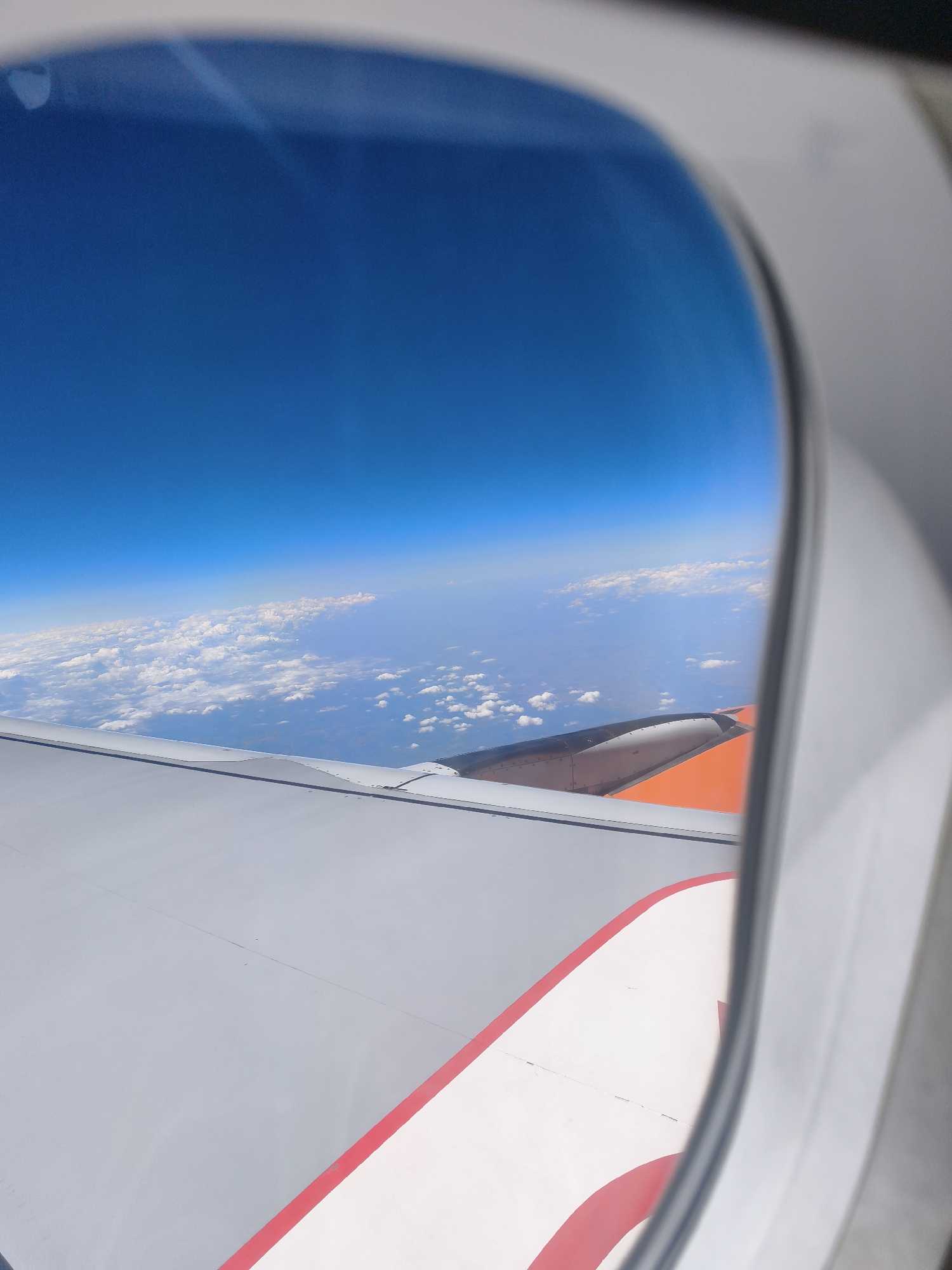 The image is taken from an airplane window, looking out at the sky and clouds. The airplane's wing is visible in the foreground, with its grey and white surface and an orange section at the top. The sky is a clear, bright blue, with a line of white clouds stretching across the horizon. The scene does not provide any specific details or context that would allow for the identification of the city of Marchegg, Austria, or any other specific location. It is a generic aerial view from an aircraft.