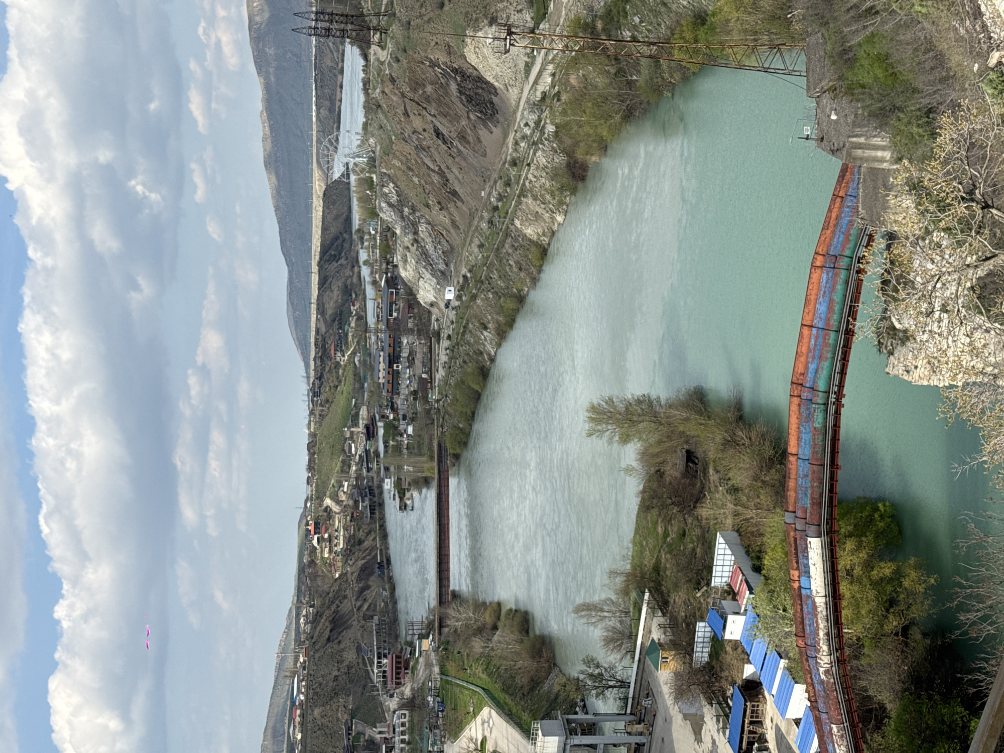 The image presents an elevated view of the city of Novo-Zubutli, Russia. A wide, light turquoise river dominates the scene, flowing through a valley. On the left bank of the river, a densely packed village extends up the slope, featuring a mix of residential buildings, structures under construction, and roads. A simple bridge spans the river in the distance. On the right bank, the terrain is characterized by rocky cliffs and sparse vegetation. A rusted pipe bridge runs along the right side of the image. Several structures with blue roofs are visible near the pipe bridge. Power lines traverse the landscape, adding vertical elements to the composition. The sky is partly cloudy.