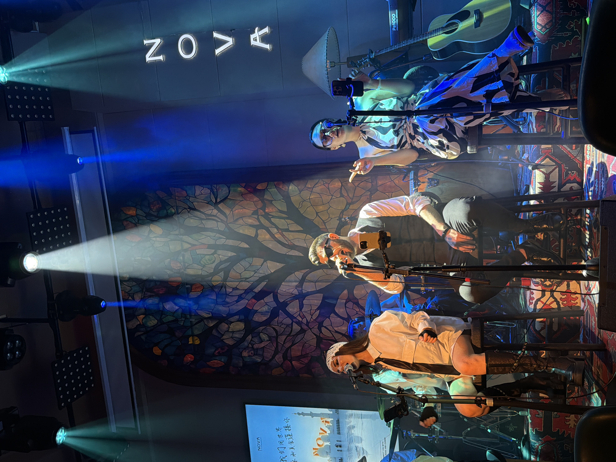 A band performs on a stage in a dimly lit venue with vibrant blue and green spotlights. The backdrop features a large, colorful stained-glass-like panel depicting a tree with various colored leaves. In the foreground, three musicians are seated on stools. The leftmost musician, a woman, wears a white collared shirt, black tie, and black thigh-high boots. She sings into a microphone, her eyes closed. To her right, a man in a grey vest over a white shirt and dark pants, wearing sunglasses, also sings into a microphone. He holds a smartphone on a stand. The rightmost performer, a woman, wears a black and white patterned dress and glasses, and gestures with her left hand while singing into a microphone and holding a smartphone. Behind the performers, musical instruments including drums, a guitar, and a keyboard are visible. The word "NOVA" is illuminated in large white letters on the dark wall behind them. A screen displaying text and an image of buildings and a sunset is visible to the left. The stage is adorned with a patterned rug. The scene appears to be in Yuzhong District, China.