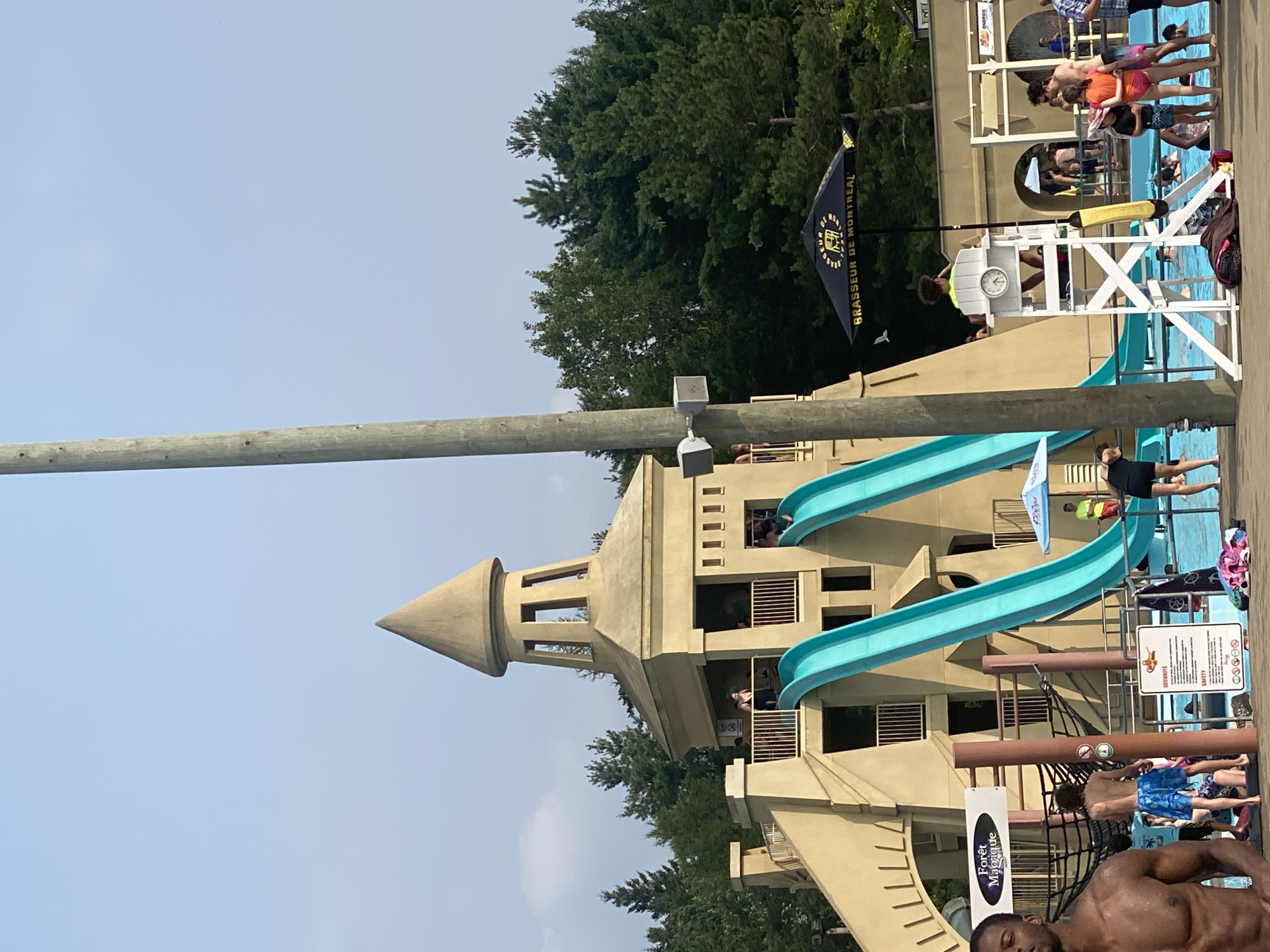 The image depicts a water park with a large, tan-colored structure resembling a castle, featuring two prominent turquoise water slides. The structure has multiple levels with balconies and arched openings, and a conical tower on top. People are visible on the balconies and near the top of the slides, with some appearing to be queuing. A lifeguard is seated in a high chair near a swimming pool, which is filled with people. Other water park amenities, such as umbrellas, lounge chairs, and signs, are present. The background consists of a dense line of green trees under a clear blue sky. The man in the foreground on the left appears to be a patron. The location is identified as being in Saint-Gabriel-de-Valcartier, Canada.