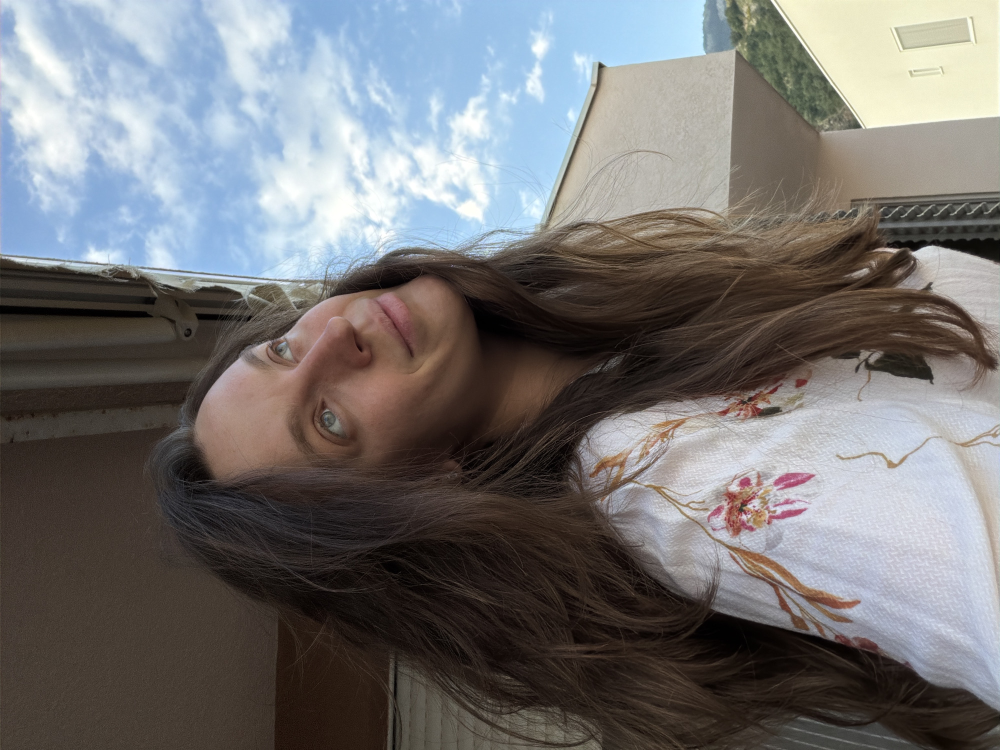 A woman with long brown hair and green eyes is looking at the camera. She is wearing a white shirt with a floral print. The background shows a blue sky with clouds and the upper part of a building with mountains in the distance. The location is Budva, Montenegro.