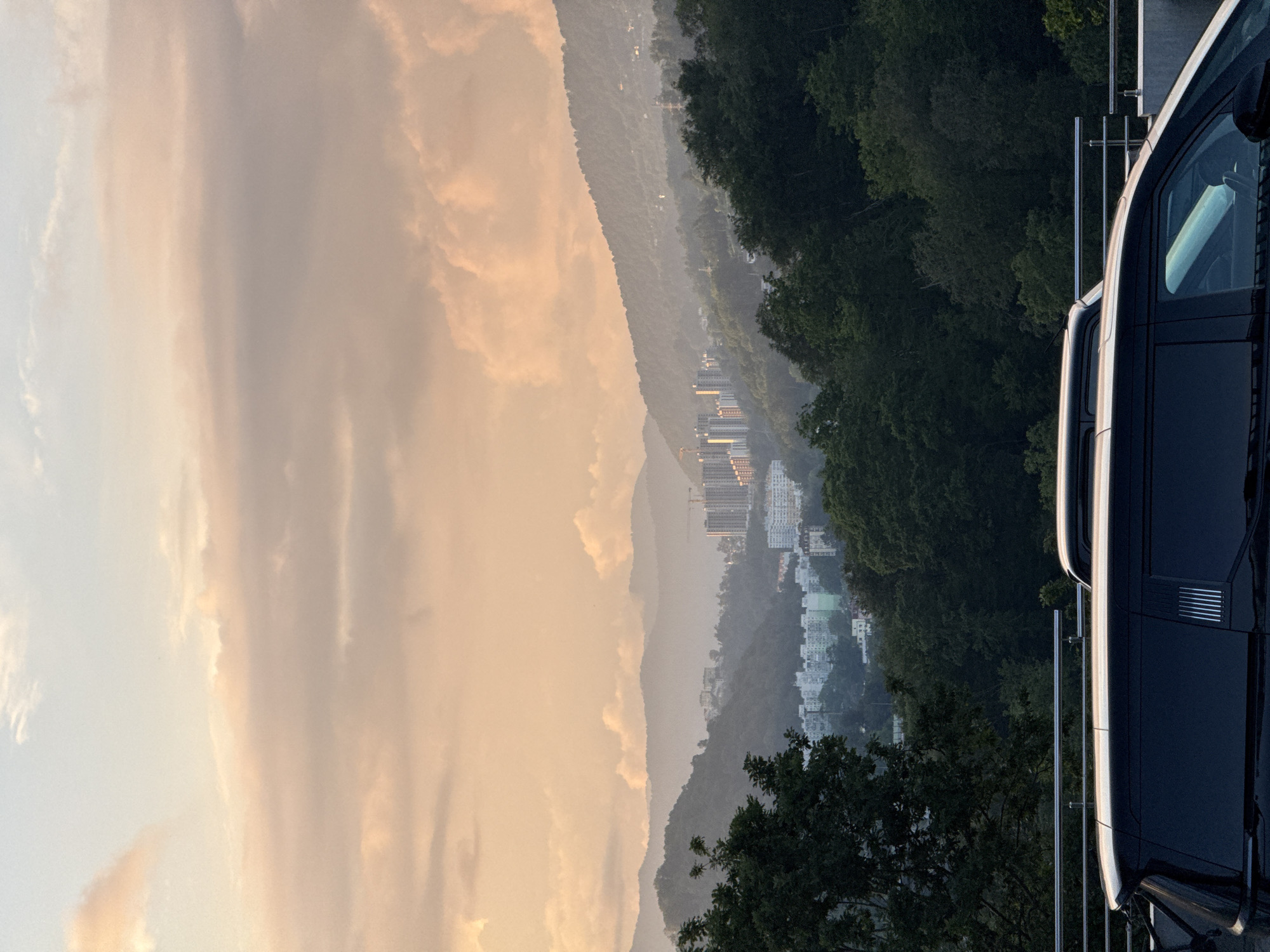 The image captures a scenic view of a city nestled among rolling hills, possibly in or around Sochi, Russia, during what appears to be sunset or sunrise. The sky is filled with soft, diffused light, casting a warm, peachy glow on the scattered clouds. In the mid-ground, several multi-story buildings are visible, some appearing to be residential complexes, dotting the hillsides. A few construction cranes are also present, indicating ongoing development. Dense green foliage covers the lower slopes of the hills, transitioning into the built-up areas. In the foreground, the top portions of two dark-colored vehicles, likely SUVs or vans, are visible. They are parked alongside a metal railing, suggesting a viewing area or a roadside overlook. The overall scene conveys a sense of tranquil urban life integrated with natural mountainous landscapes.