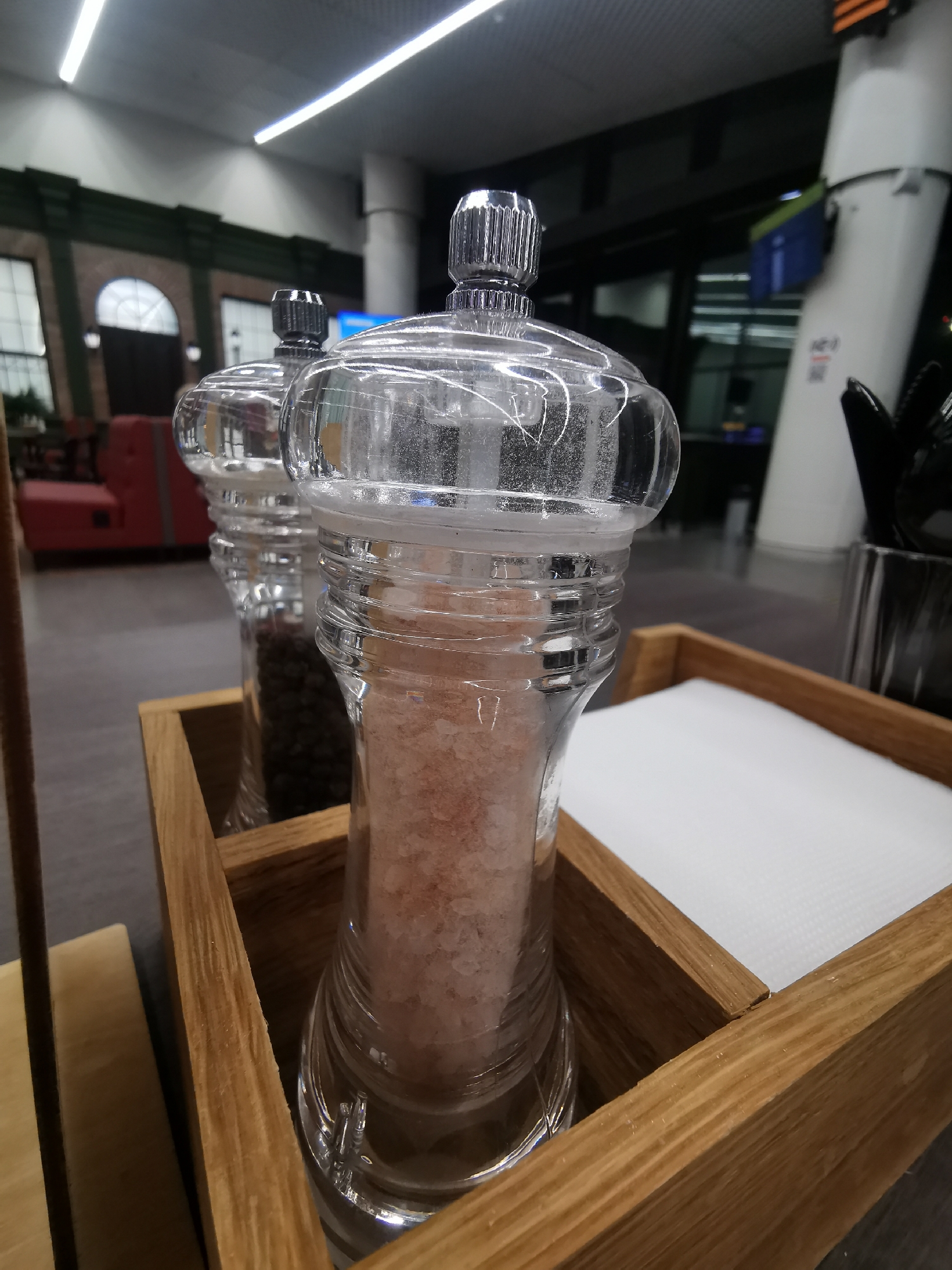 In a well-lit indoor setting, likely a restaurant or dining area, two transparent grinders stand side-by-side in a wooden tray. The grinder in the foreground is filled with pink Himalayan salt, while the one behind it contains black peppercorns. Adjacent to the grinders in the same tray, a stack of white napkins sits neatly. The grinders feature clear glass bodies with a ridged design and a metal cap on top.
The tray is made of light-colored wood, adding a rustic touch to the setup. In the background, there is a glimpse of a seating area with a red couch and a window. Further in the background, a tall white structure is visible, potentially part of the building's architecture or a decorative element. The environment suggests a modern interior with a blend of natural and artificial lighting.
