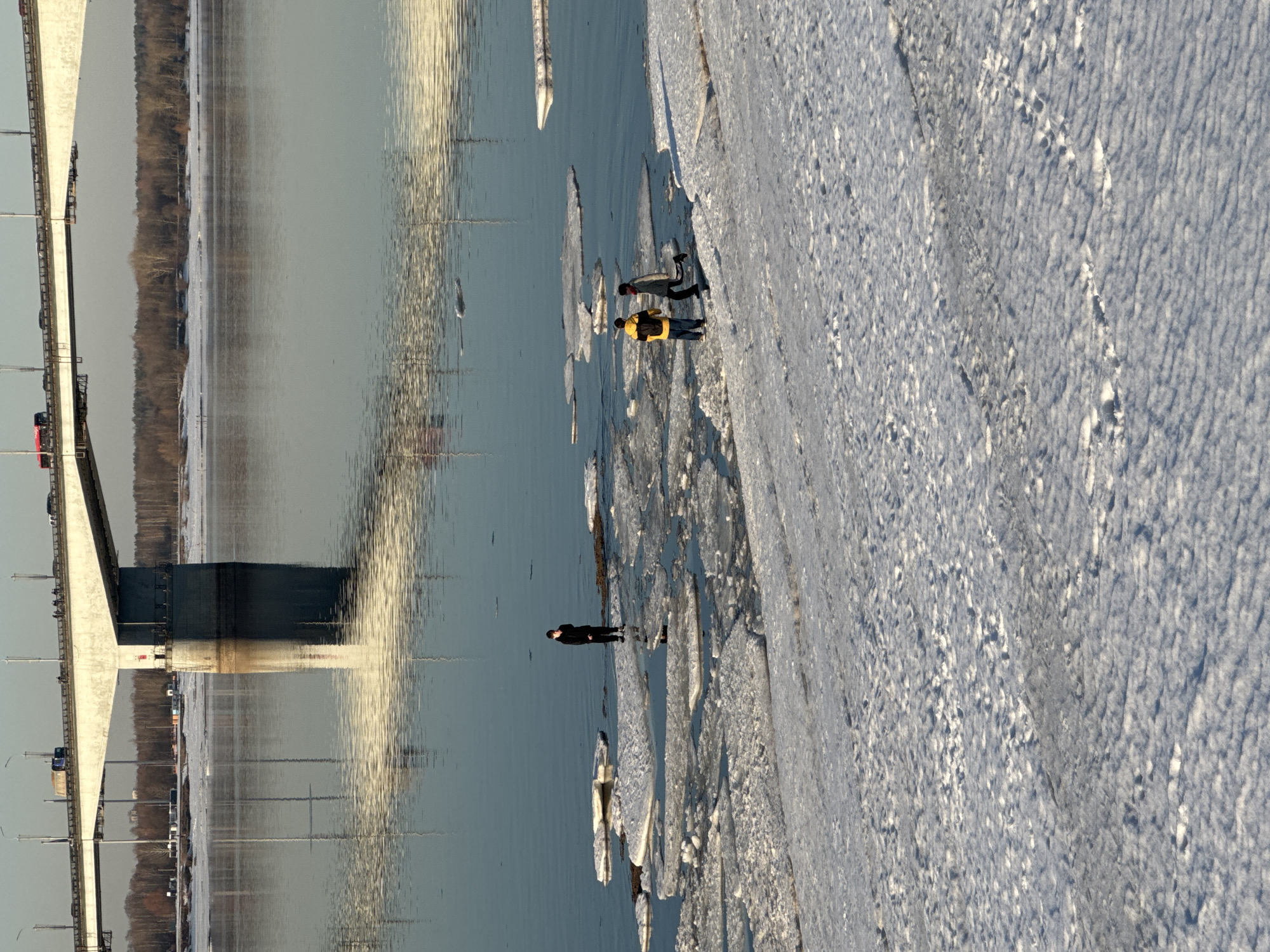 The image depicts a winter or early spring scene along the Kama River in Perm, Russia, with the Kommunalny Bridge prominent in the background. The foreground shows a snow-covered riverbank with numerous visible footprints. Three individuals are situated near the water's edge. On the left, a single person in dark clothing stands on a small isolated section of ice or land. To the right, two individuals are present: one wearing a yellow jacket and a dark backpack stands on the snow-covered bank, while the other, in dark clothing with a red neck covering, is in motion, appearing to step onto an ice floe. The river surface contains scattered, irregular ice floes and reflects the overhead bridge and sky. In the upper background, the Kommunalny Bridge, a large concrete structure, carries vehicular traffic, including a red bus and a blue truck, across the river. The distant shoreline beyond the bridge is lined with leafless deciduous trees and patches of snow. The sky is a uniform light gray, and strong reflections on the water indicate bright, diffused light.