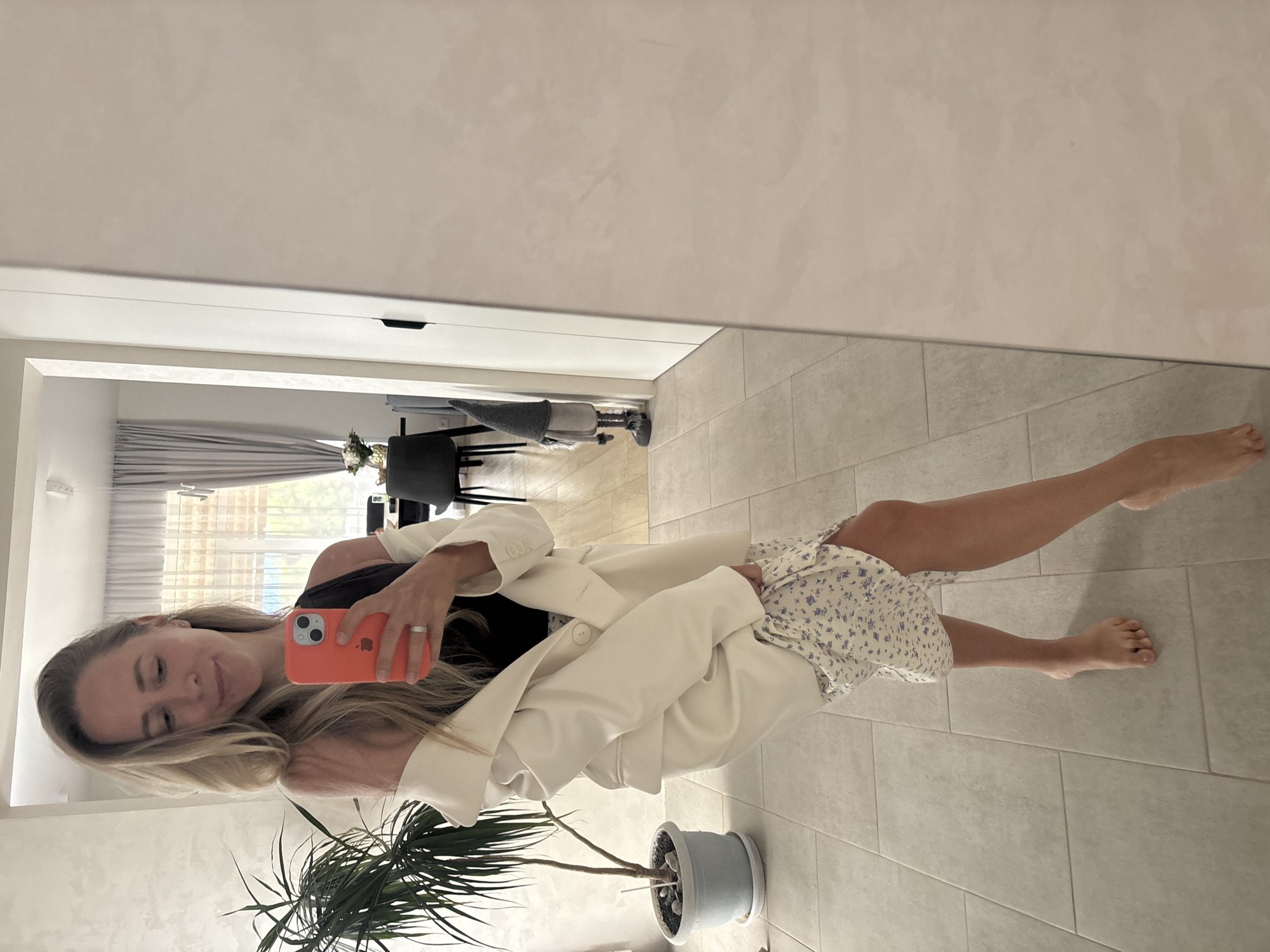 A woman stands in a room, holding a phone in her right hand and looking down at it. She is wearing a white blazer over a dark top and a floral-print skirt. Her left hand is lifting the hem of the skirt. She is barefoot. In the background, there is a plant, a doorway, and a mirror. The room appears to be a residential interior, possibly an apartment. The scene is well-lit with natural light. The location is not identifiable as Chaikovsky, Russia.