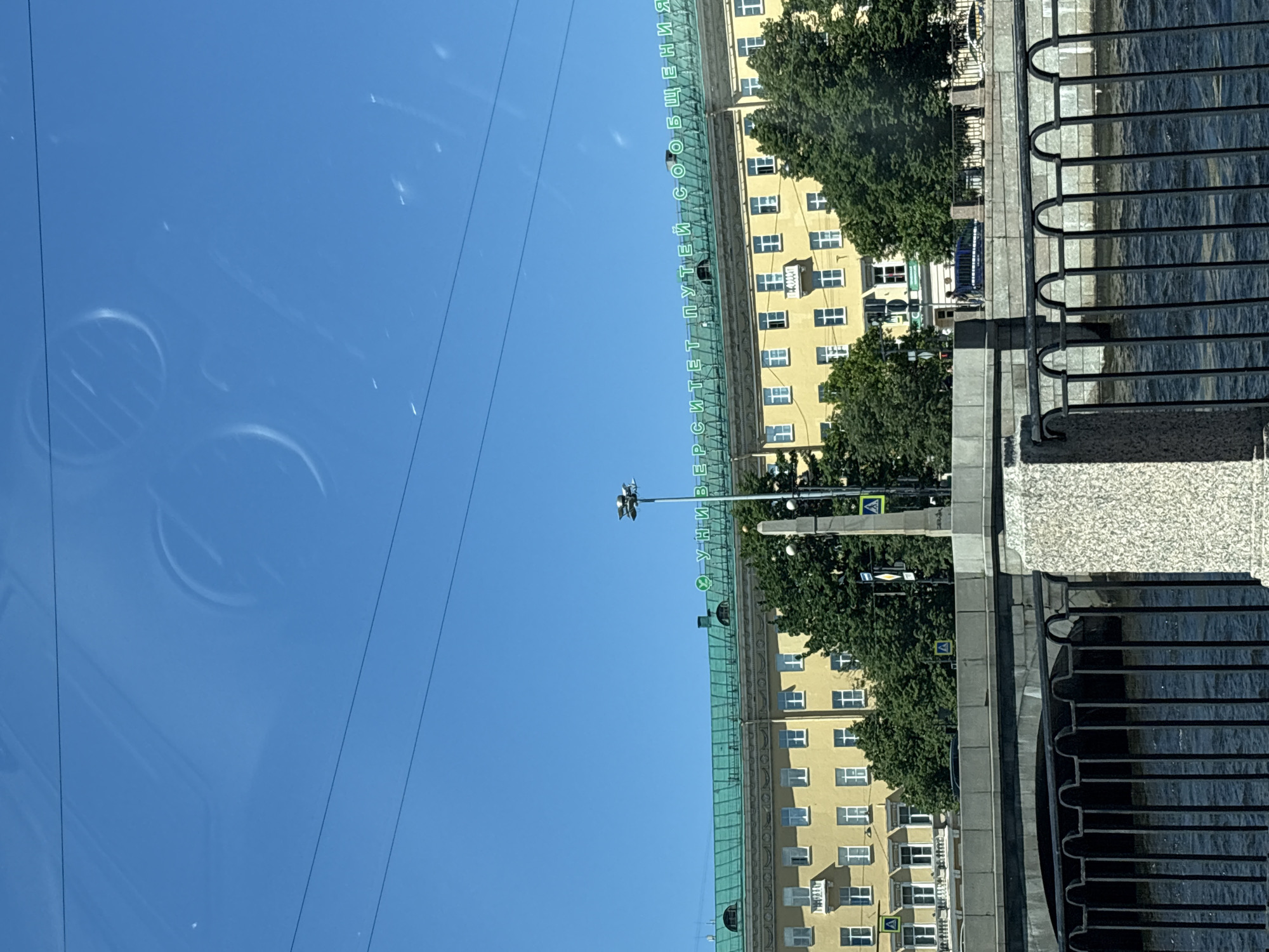 The image shows a building with green lettering on its roof that reads "УНИВЕРСИТЕТ ПУТЕЙ СООБЩЕНИЯ" which translates to "University of Communications." The building is yellow with a green roof and many windows. It is located in Saint Petersburg, Russia. A bridge, a body of water, and trees are also visible in the image. The sky is clear and blue.