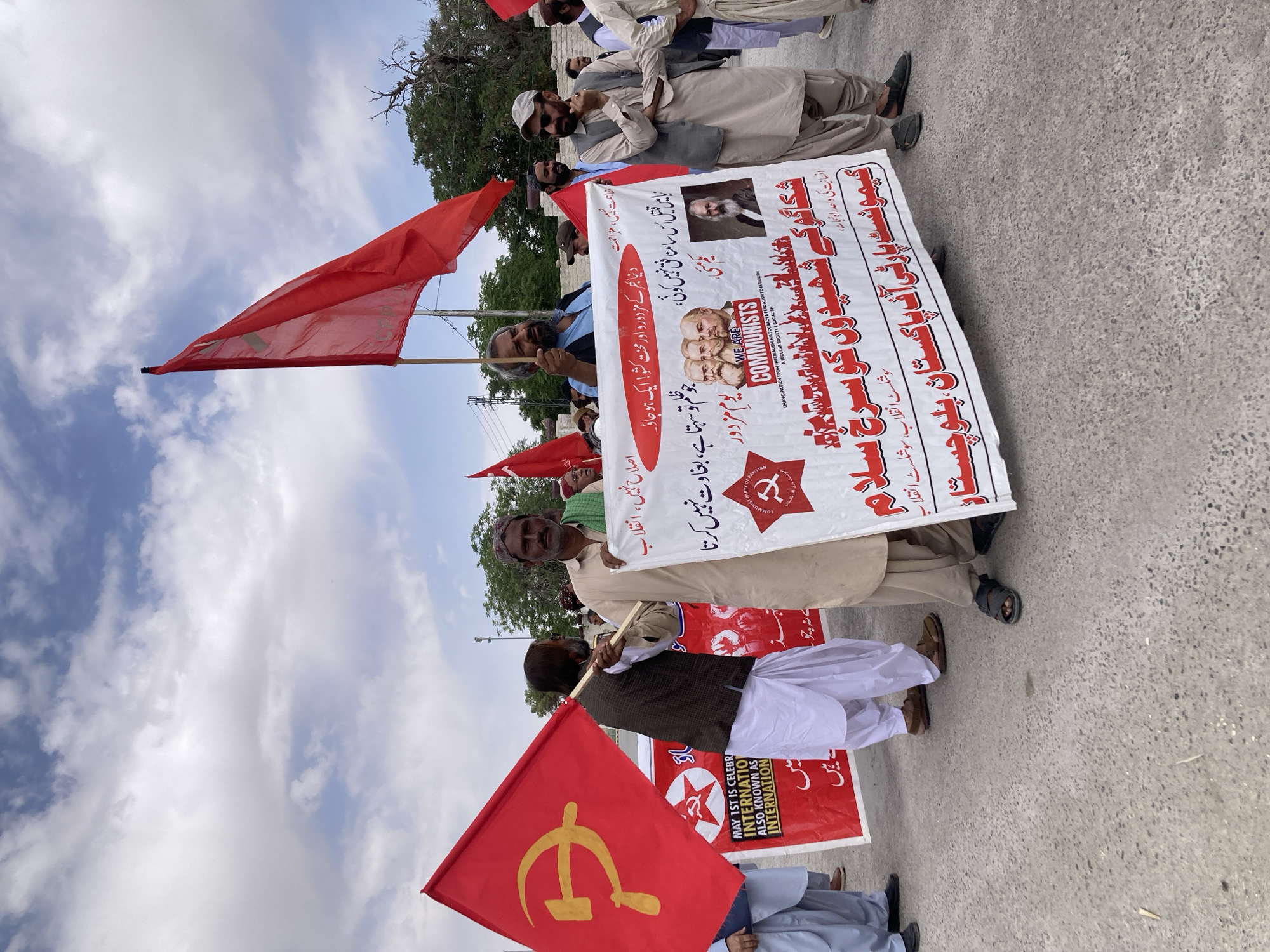 This image captures an outdoor political rally or demonstration held on a paved road or open ground in Quetta City Tehsil, Pakistan. A group of men, dressed in traditional South Asian attire including shalwar kameez and waistcoats, are participating in the event.
The central elements of the scene are the multiple red flags and large white banners. Many flags feature communist symbols such as the hammer and sickle, and a red star enclosing a hammer and sickle. The primary white banner, held prominently by several participants, displays portraits of historical communist figures, specifically Karl Marx, Friedrich Engels, and Vladimir Lenin.
The banners and signs contain both English and Urdu text. English phrases include: "WE ARE COMMUNISTS," and "EMANCIPATION FROM IMPERIALISM, HILTOCRACY & FEUDALISH TO ESTABLISH A SECULAR SOCIETY & SOCIALISM." Visible Urdu text translates to: "Red salute to the martyrs of Chicago," "The only path to salvation for humanity," "Socialist Revolution, Socialist Revolution," and identifies the group as "Communist Party of Pakistan, Balochistan." Other slogans present are "May 1st," "Workers and laborers of the world! Unite!", "No reform, revolution," and a quote: "There is no hypocrite like Qatil in the law who tolerates oppression but does not rebel." A smaller flag also makes reference to "MAY 1ST IS CELEBRA... INTERNATIO... ALSO KNOWN AS INTERNATION..." suggesting a May Day celebration.
The setting is outdoors during daytime, under a partly cloudy sky. Trees and what appear to be utility poles are visible in the background, indicating an urban or semi-urban environment. The overall activity is a focused political gathering promoting communist ideology and workers' rights.