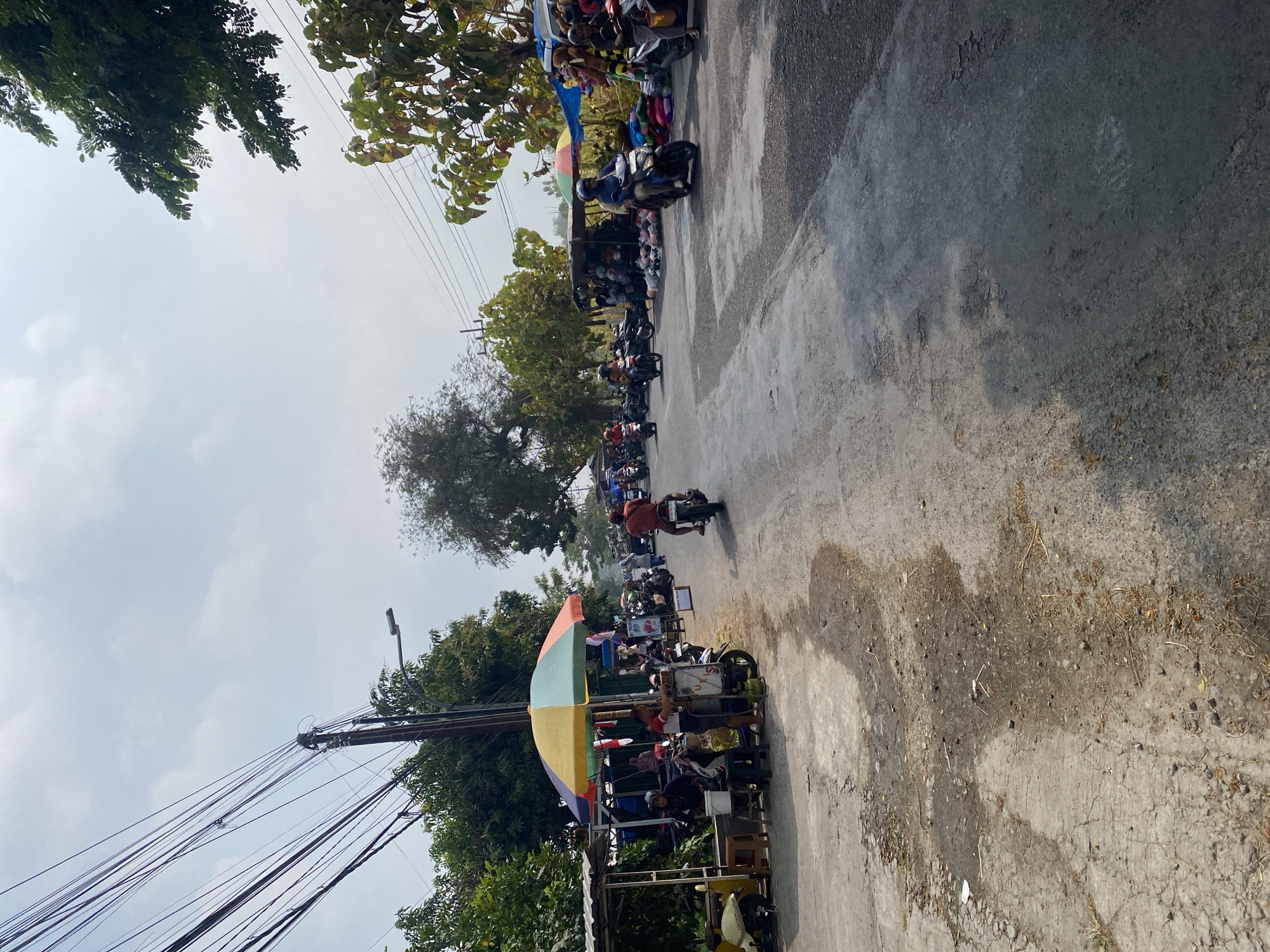 The image depicts a street scene in Sempol, Indonesia, characterized by a dusty, worn asphalt road lined with temporary market stalls and various vehicles. The sky is overcast, suggesting a warm, potentially humid day.
On the left side of the road, a market stall is set up under a colorful umbrella, with several people gathered around. A scooter is parked to the far left. The stall appears to be selling food or drinks, as indicated by the presence of a gas cylinder and what looks like seating. Further down the street, more stalls are visible, draped with blue tarps, displaying goods such as stuffed animals and other merchandise.
The road itself is busy with motorbikes, the primary mode of transportation. Several individuals are seen riding motorbikes, navigating through the market area. Some are heading towards the viewer, while others are moving away. The road surface is uneven and shows signs of wear and tear, with patches of dirt and debris.
In the background, trees and power lines are prominent features of the environment. The power lines are numerous and appear to be strung between wooden poles, some of which are leaning. The vegetation is lush, indicating a tropical or subtropical climate. The overall impression is one of a lively, informal marketplace in a rural or semi-urban setting.