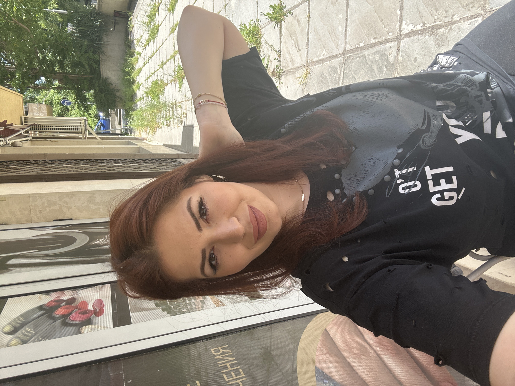 A woman is posing for a photo outdoors in Sofia, Bulgaria. The woman has auburn hair and is wearing a black t-shirt. She is smiling and holding one arm up. The background includes a sidewalk, building facade, greenery, and street. The image also captures a storefront window displaying images of nail polish and manicured hands, as well as text in a Cyrillic script.