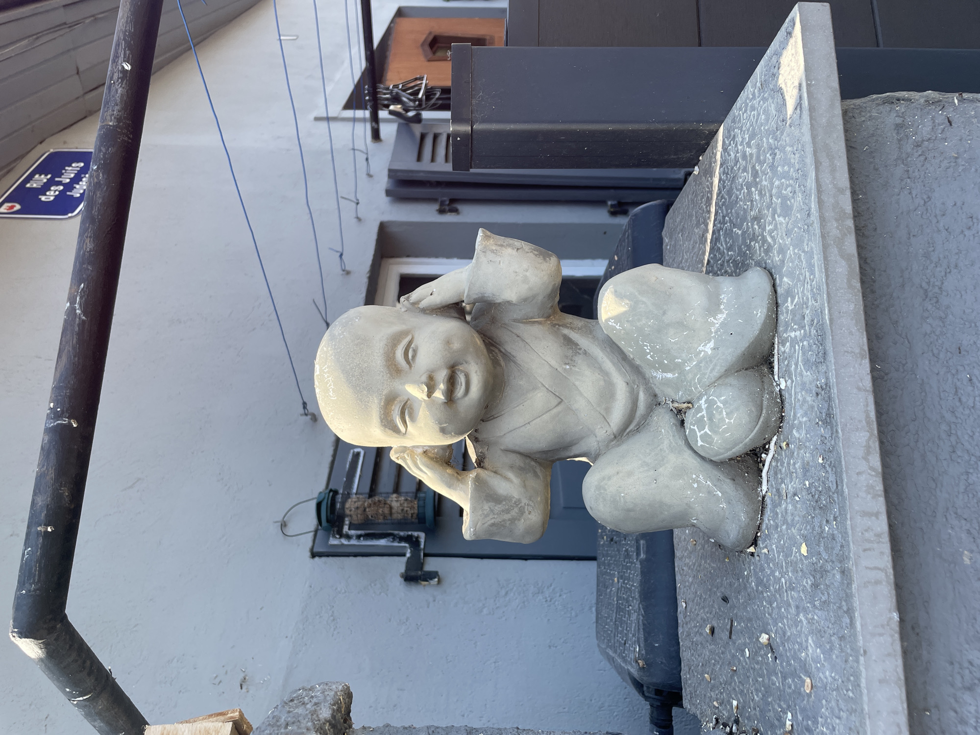 A close-up, rotated view shows a light gray stone statue of a baby in a relaxed pose. The statue is positioned on a textured gray stone surface, with a dark gray structure behind it. The baby statue has its legs bent and its arms raised, with hands near its head. The baby's face is slightly turned, with its mouth open as if speaking or laughing. The statue has a weathered appearance with visible cracks and debris on its surface.
In the background, a light blue sign with white text is visible on a dark pole. The text on the sign reads "RUE des Juifs". Above this, a building with a dark awning and windows can be seen. A black pipe runs diagonally across the left side of the image. The scene appears to be outdoors, possibly on a balcony or ledge, with the sky suggested by the lighting. The time of day is not explicitly clear, but the lighting suggests it might be daytime. The weather appears to be clear, with no visible rain or clouds. The location context is Gerstheim, France, with the street name providing a specific clue to the location.