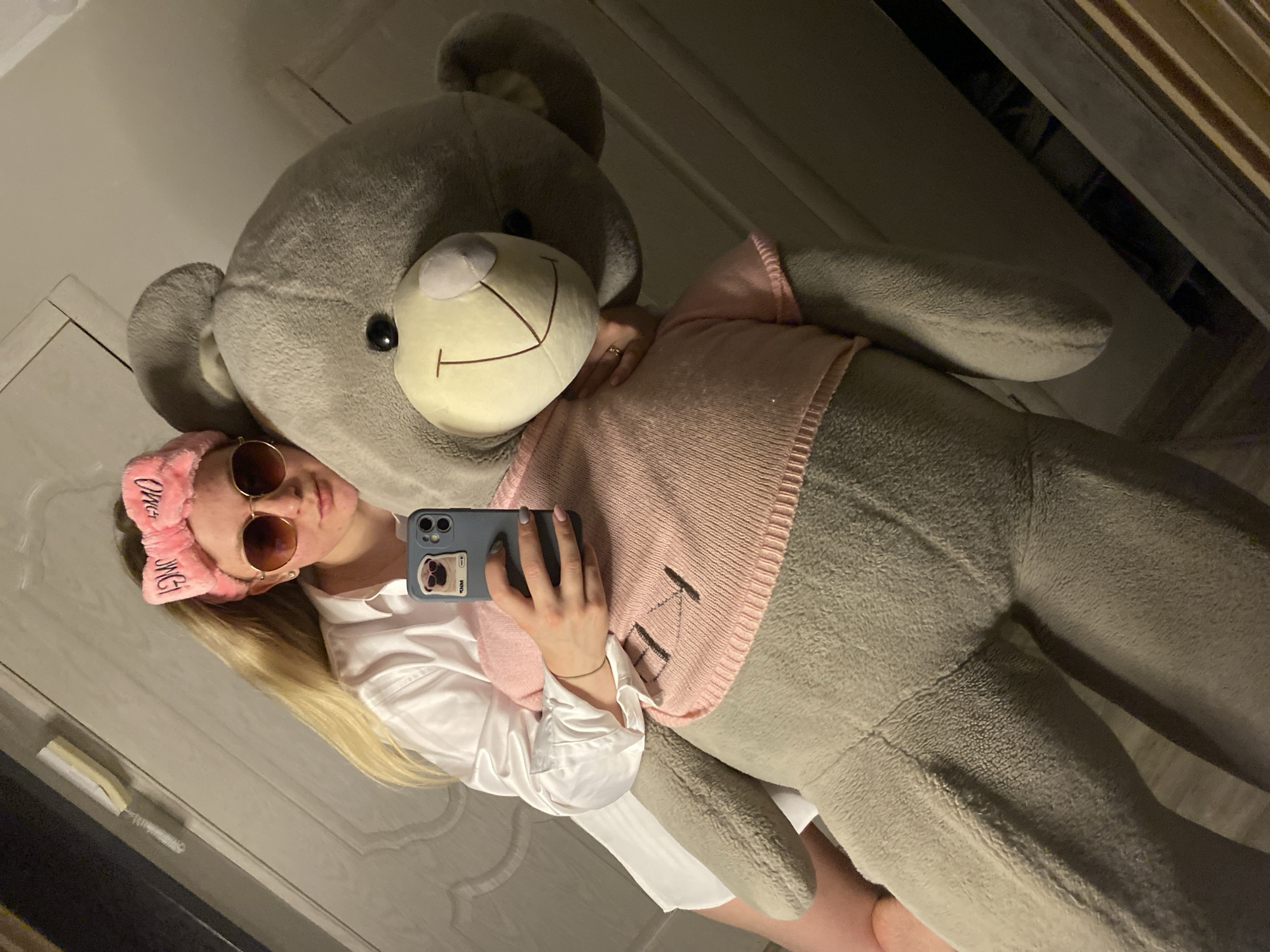 An indoor, mirror-reflected full-length photograph captures a blonde individual, appearing to be a young woman, embracing a large grey teddy bear. The woman is wearing a white button-up shirt, oversized round sunglasses, and a pink fluffy headband with "OMG" printed on it. She holds a smartphone with a light blue case featuring a pug in sunglasses, angled towards the mirror to take the photograph. Her left hand is visible, adorned with a ring and light pink nail polish. The teddy bear, approximately the same height as the woman, is clad in a pink knitted sweater with a dark stitched design on the chest. The bear has a black nose and a stitched smile on its cream-colored muzzle. The background reflects parts of an interior room, including a white door or wall paneling and a narrow, light-colored object on an upper ledge. The floor appears dark. The scene takes place in a location in the city of Unknown, Unknown.