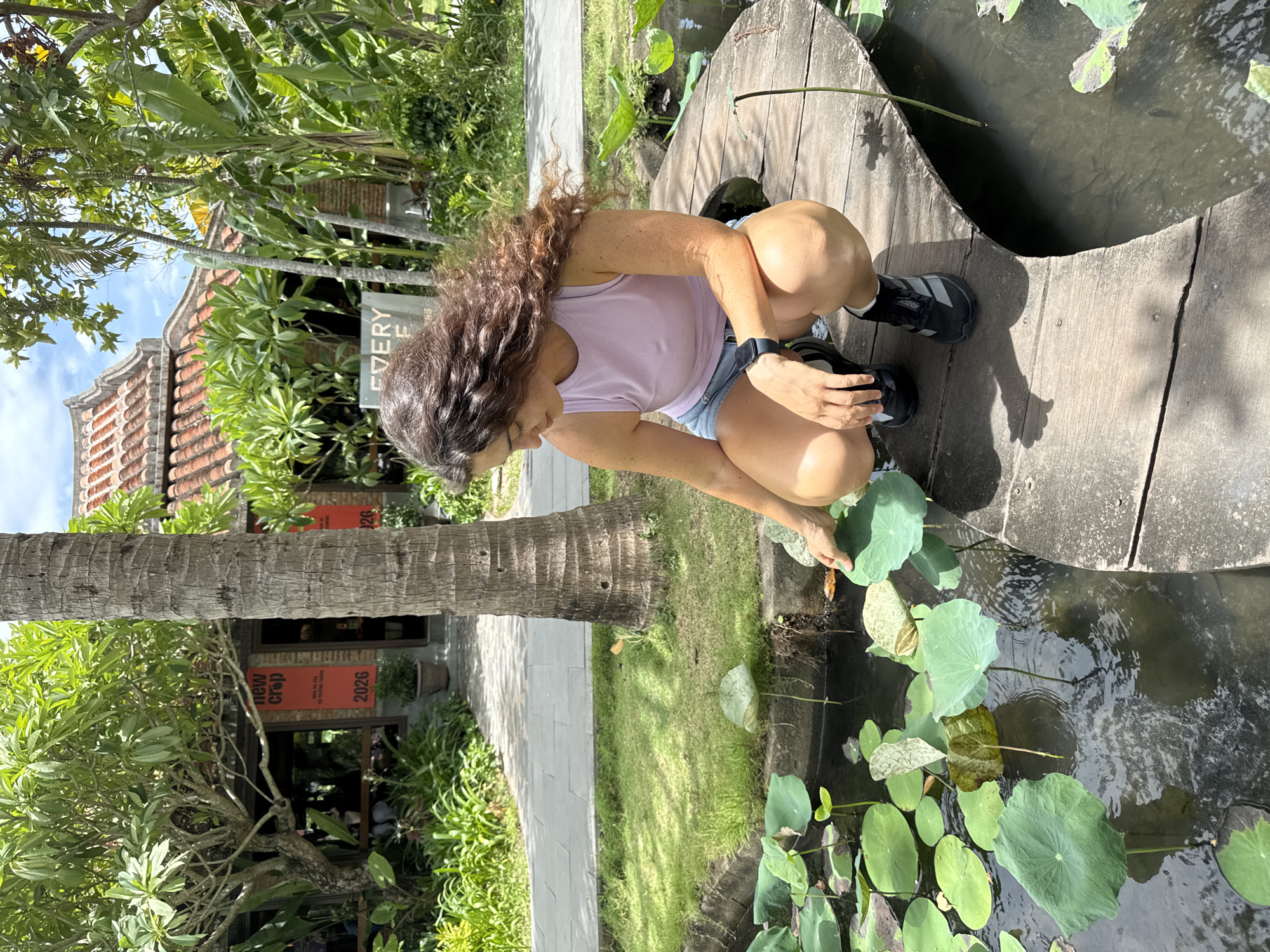 A woman with long, dark curly hair, wearing a pale pink tank top and denim shorts, is crouching on a wooden walkway that curves over a pond filled with lotus pads. Her right hand is extended, touching a lotus pad. She is wearing black sneakers with white accents and a black smartwatch on her left wrist. The walkway is made of weathered wooden planks. Behind her, a tall palm tree trunk stands vertically. In the background, a building with a red-tiled, traditional Vietnamese roof is visible, surrounded by lush green foliage, including banana trees and smaller leafy plants. A red banner with "new crop" and "2026" printed on it hangs from the building. The scene appears to be in Hoi An, Vietnam, likely at a cafe or restaurant given the signage. The lighting suggests it is daytime, with a partly cloudy sky.