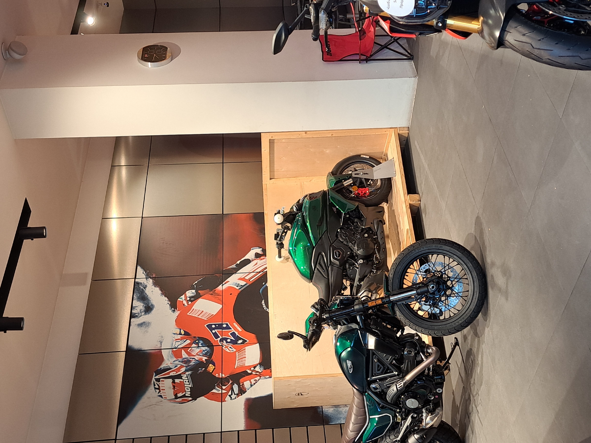 The scene depicts a showroom in Yerevan, Armenia, featuring a Ducati motorcycle display. Two green Ducati motorcycles, likely Scrambler models, are prominently positioned. One is partially encased in a wooden crate, while the other is freely displayed on the showroom floor. A large mural depicting a motorcycle racer, likely a Ducati team member, adorns the wall behind the motorcycles. The showroom has a modern aesthetic, with neutral-toned walls and track lighting. Another Ducati motorcycle is visible in the background, along with promotional materials.