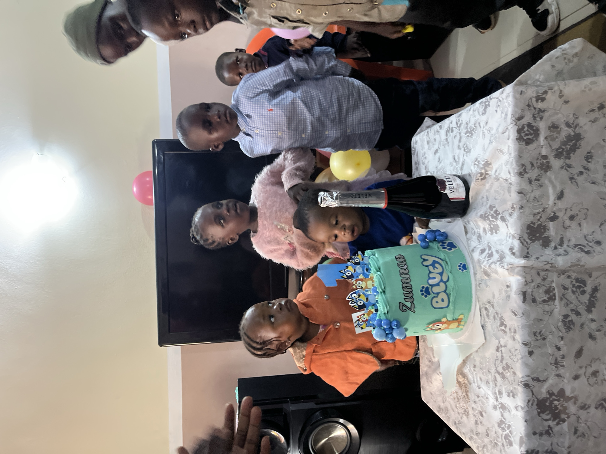 The image captures a birthday celebration for a child named Zunnan, whose name is written on a turquoise cake decorated with Bluey characters and a large number "1". The cake sits on a table covered with a white, floral-patterned tablecloth. A bottle of "VELETA" champagne is also on the table. Several children and at least one adult are present, gathered around the table. The children are of varying ages, with some looking towards the cake and others looking off-camera. One young child in an orange jacket stands closest to the cake. In the background, a television screen is visible, and pink and yellow balloons are scattered around the room. The setting appears to be an indoor residential space in Bukuru, Nigeria, indicated by the overall decor and the presence of a television and domestic furnishings. The lighting is artificial, contributing to a warm, celebratory atmosphere.