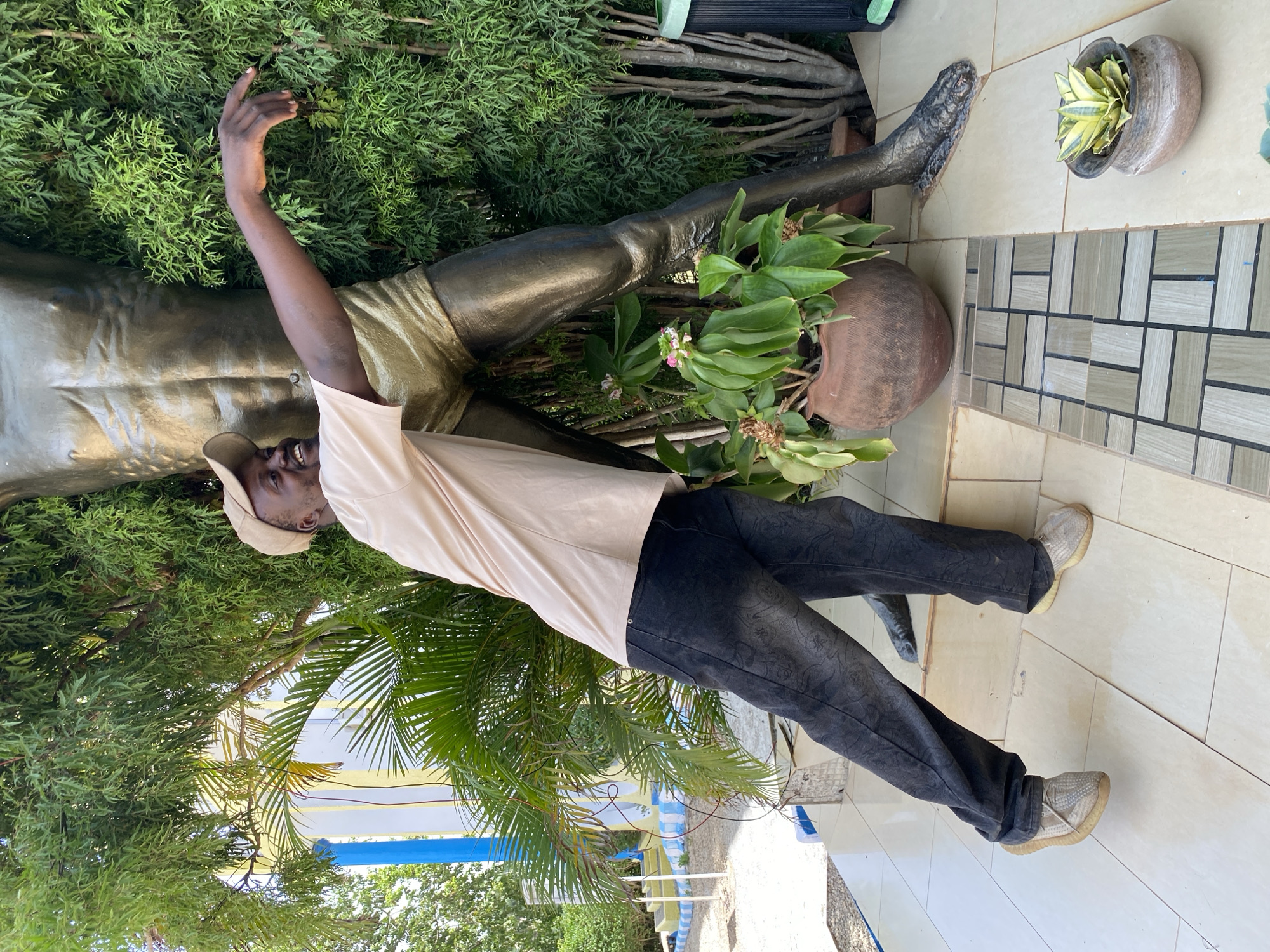 A man stands next to a bronze statue of a nude male figure on a tiled surface in what appears to be an outdoor setting. The man wears a tan baseball cap, a light beige t-shirt, dark pants with a patterned design, and light-colored sneakers. He has one arm raised toward the statue's hand. The statue appears to be resting on or partially obscured by various plants. Other plants, including potted ones, are in the background, and a palm tree is visible. The location is Barakin Angundi, Nigeria.