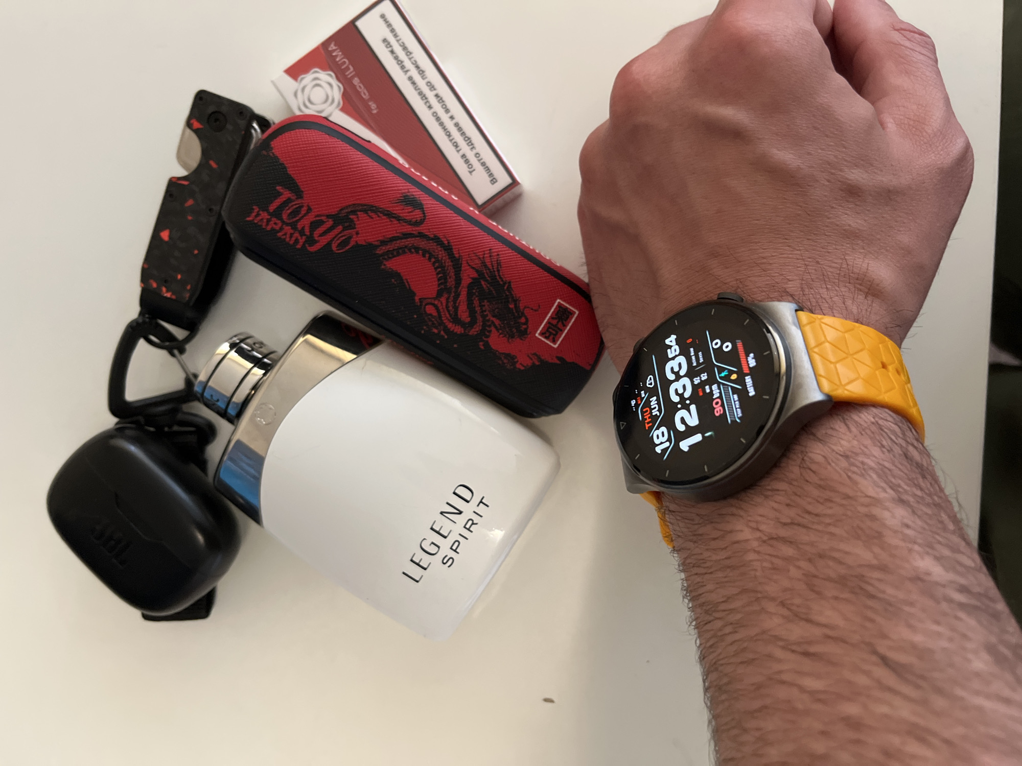 The image shows a flat lay of personal items on a light-colored surface, likely a table. The objects include a black JBL earbud case, a white bottle of "LEGEND SPIRIT" perfume, a small multi-tool with a red and black speckled handle, a black and red pouch with a dragon design, and a red and white packet labeled "for IQOS ILUMA" with Cyrillic text. A smartwatch with a bright yellow band is worn on a person's hairy wrist, displaying the time as 12:33, the date as Thursday, June 18th, and various health metrics. The background appears to be a neutral, indoor setting with a white surface, possibly a desk or countertop. There is no indication of the city of Sofia, Bulgaria, within the image itself.