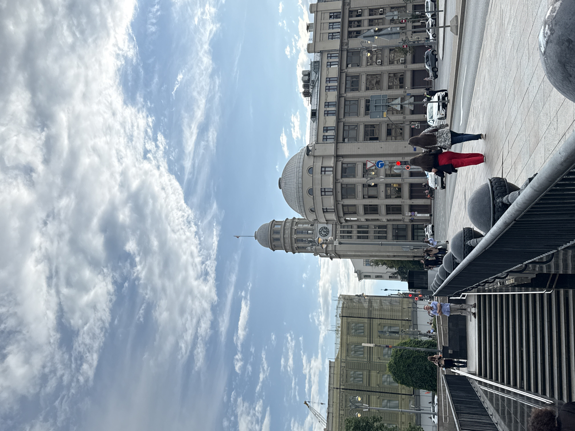 This is a medium shot, taken from a slightly elevated perspective, of a street scene in Moscow, Russia, possibly near the Red October confectionery factory, given the architectural style. The sky is a mix of blue and white clouds. In the foreground, a set of concrete stairs with metal railings leads down to a street level. A man in a light blue shirt and khaki pants stands at the top of the stairs with his arms crossed, facing away from the camera. Further down the street, two women in red pants and a patterned jacket are walking away from the camera. They are walking on a sidewalk next to a road with several cars. On the right side of the street, there are large, ornate buildings with many windows. One building has a domed roof, and another has a clock tower. On the left side of the street, another building is under construction, with scaffolding and a crane visible. Streetlights are present throughout the scene. The overall atmosphere is that of a busy urban environment.