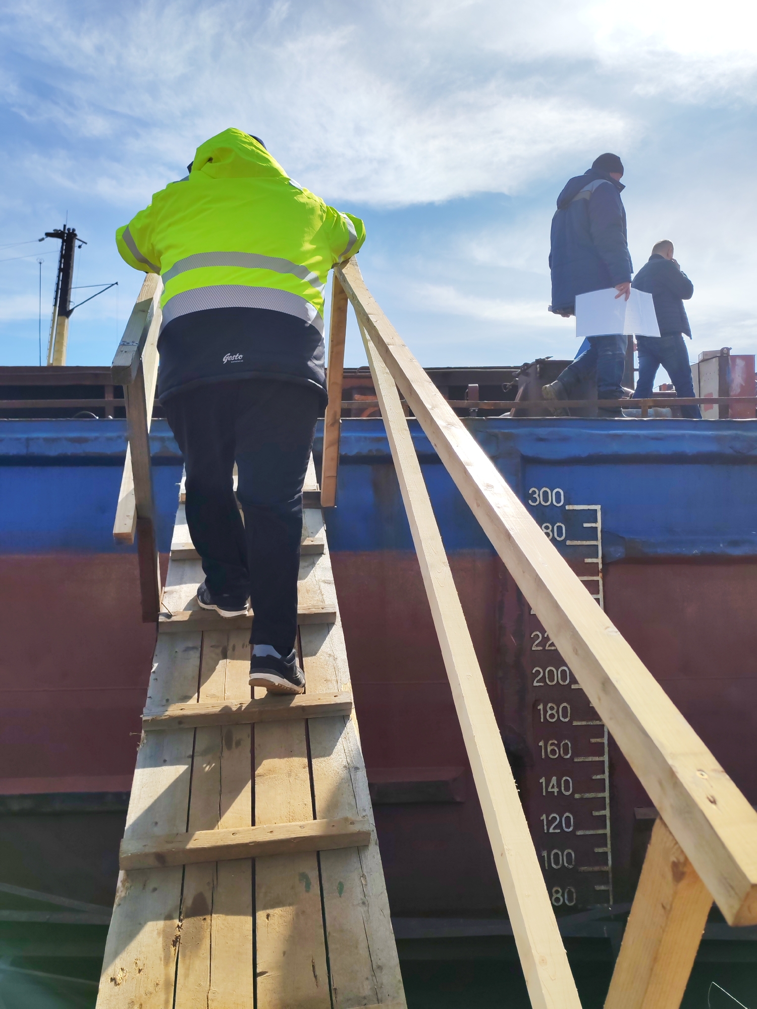 The image shows a person wearing a bright yellow jacket and dark pants climbing a wooden ladder onto a large blue and red cargo ship. The person is seen from the back, ascending the ladder.
On the deck of the ship, two other individuals are visible. One person is wearing a dark jacket and holding a white document, while the other person stands nearby.
The ship has markings resembling a draft scale painted on its side, with numbers ranging from 80 to 300 visible.
The location is identified as Bol'shie Mkhi, Russia, based on the surrounding clues and elements in the image. The environment suggests a port or industrial area with the presence of a cargo ship. The sky is partly cloudy with a mix of blue and white.