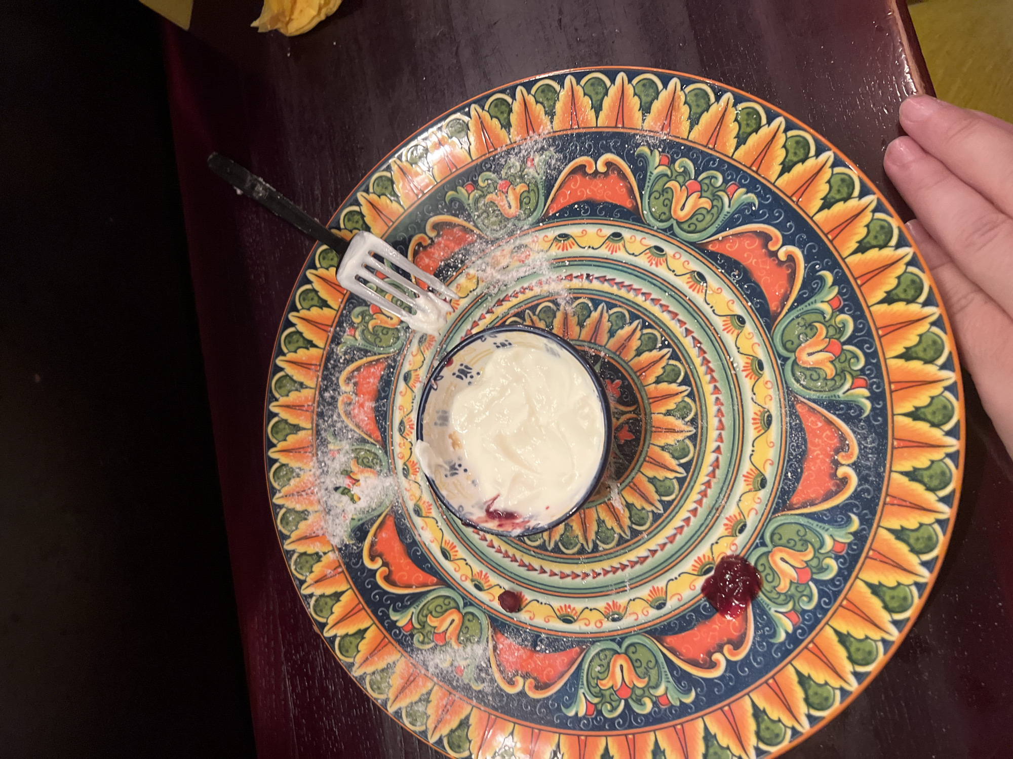 The image shows a close-up of a decorative plate with a small bowl of yogurt and a fork. The plate has an intricate, colorful pattern with floral motifs in shades of orange, yellow, green, and blue. There are specks of powdered sugar scattered on the plate. A small dollop of a dark red substance, possibly jam or fruit compote, is visible on the left side of the plate. A black fork with a white tip is resting on the plate, with some yogurt on its tines. The scene is set on a dark wooden surface, likely a table. In the upper portion of the image, a dark, out-of-focus background suggests an indoor setting. A person's hand is visible in the lower right corner, suggesting they are about to interact with the food or plate. There is no indication of the location in Saint Petersburg, Russia.