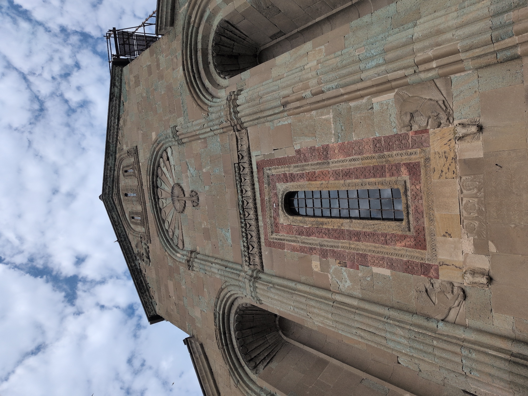 The image shows a low-angle, exterior view of a stone building, likely a church, against a cloudy sky. The architecture features arched windows, decorative carvings, and a narrow, tall window with a dark, grid-like pane in the center. The stone appears weathered and has a rough texture. Scaffolding is visible on the upper right portion of the building, suggesting ongoing construction or renovation. Inscriptions in an unknown script are visible on the lower stone blocks. The overall impression is of an ancient and imposing structure. This building is the Svetitskhoveli Cathedral, located in Mtskheta, Georgia, a UNESCO World Heritage site and one of the most important religious sites in the country.