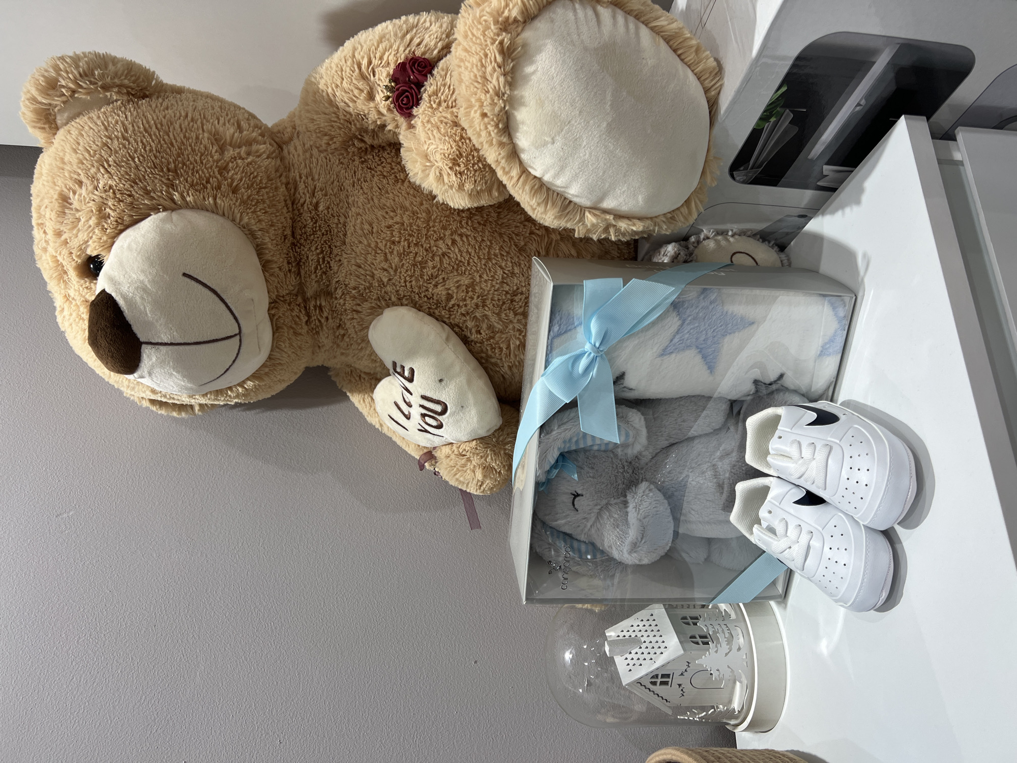 A teddy bear with a heart-shaped tag that reads "I love you" is next to a boxed gift set containing a stuffed elephant, a blanket with a blue ribbon, and a pair of white baby shoes, positioned on a white surface adjacent to a light grey wall. The surrounding environment is unknown. There is no evidence indicating that this image was taken in Gradishte, Bulgaria. No people or specific actions are visible.