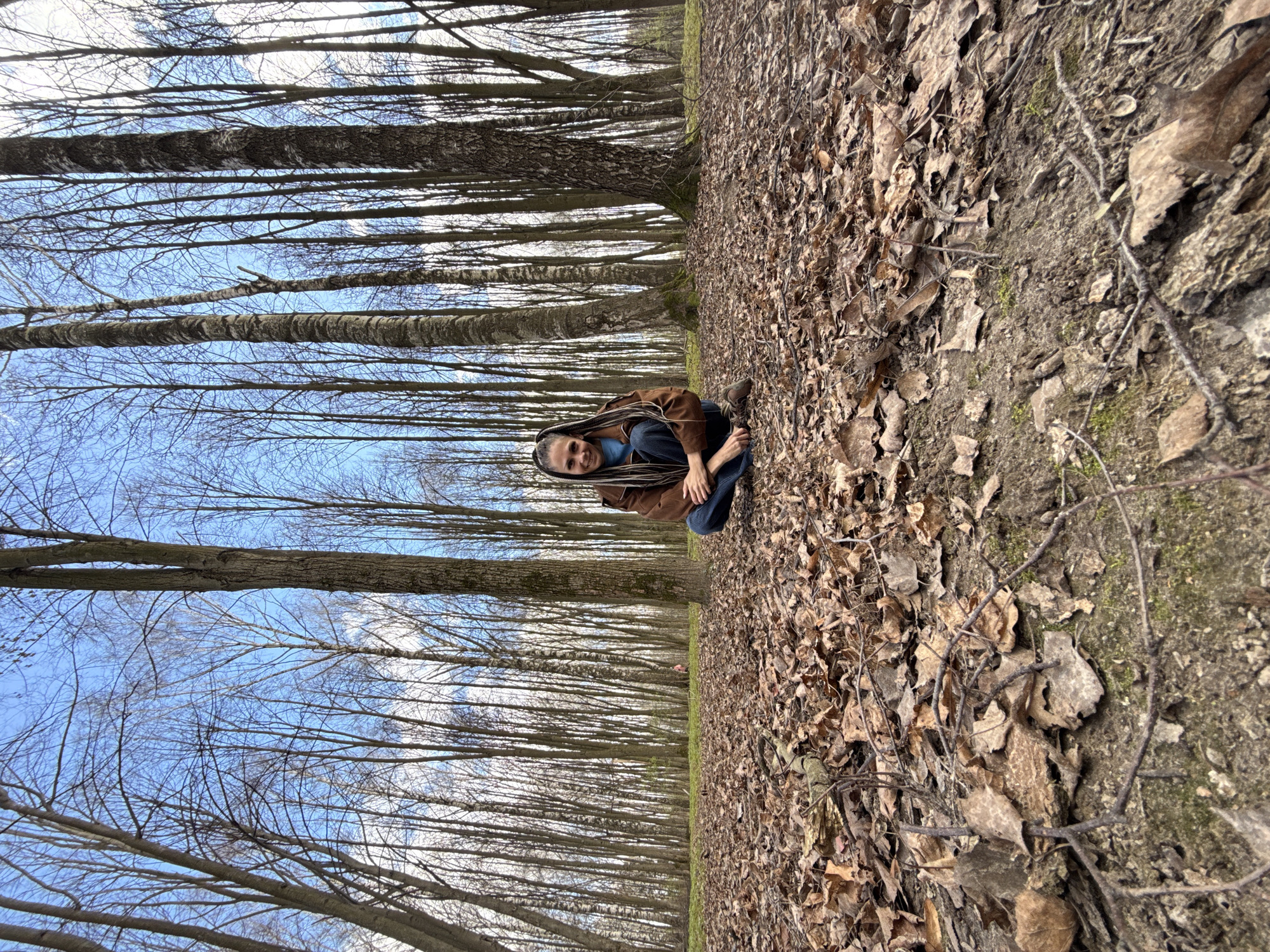 A woman with long, braided hair sits on a leaf-covered ground in a forest of tall, bare trees. She is wearing a brown jacket and blue jeans. The trees stretch into the distance under a blue sky with some clouds. The ground transitions from leaves to dirt on the right side of the frame. The image is rotated 90 degrees clockwise. The exact location in the city of Rusanovo, Russia, cannot be determined from the image.