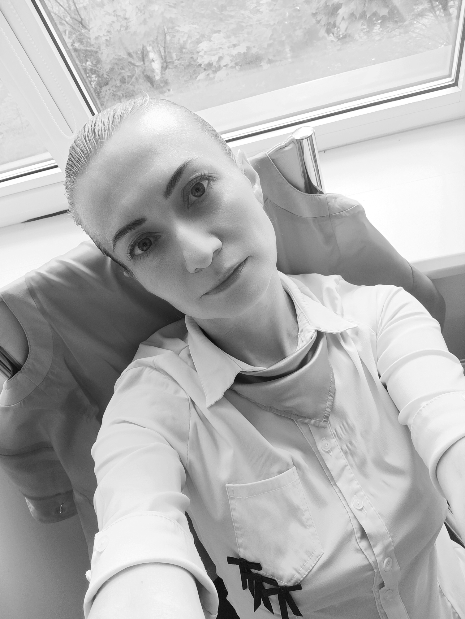A black and white close-up selfie shows a woman with short, slicked-back blonde hair and dark eyebrows. She is wearing a white collared shirt with buttons down the front and a pocket on the left side. A dark ribbon detail is visible on the lower left of the shirt. She appears to be indoors, sitting near a window with a view of trees outside, suggesting an outdoor environment visible from an urban setting. The lighting suggests it is daytime. Based on the presence of birch trees and the type of window frame, this scene could be consistent with a location in Moscow, Russia.