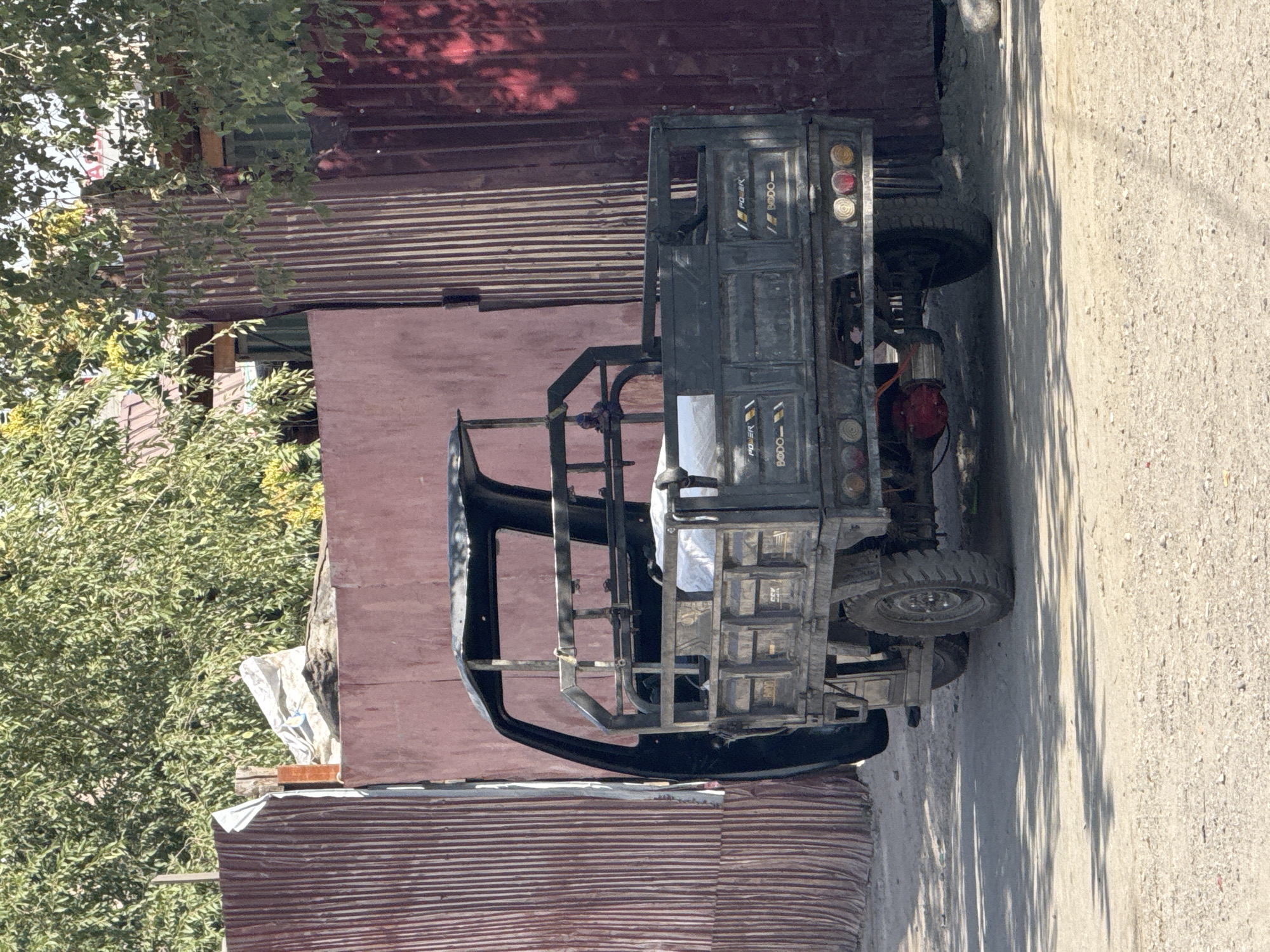 In Bishkek City, Kyrgyzstan, a three-wheeled truck is parked on a dirt road. The truck is dark gray, with a black metal frame that forms a partial cab and a rear cargo bed. The cargo bed is empty except for a white tarp or bag. The truck has two wheels visible in the back and one in the front, which is partially obscured. The rear of the truck has a red engine or transmission component and multiple circular tail lights. The truck is parked next to a corrugated metal fence that is dark red on the right and a lighter, possibly faded red on the left. Behind the fence, green trees with visible leaves are present, suggesting an outdoor, possibly residential or industrial area. The ground is uneven and covered with gravel and dirt. Sunlight casts shadows on the road and the truck.