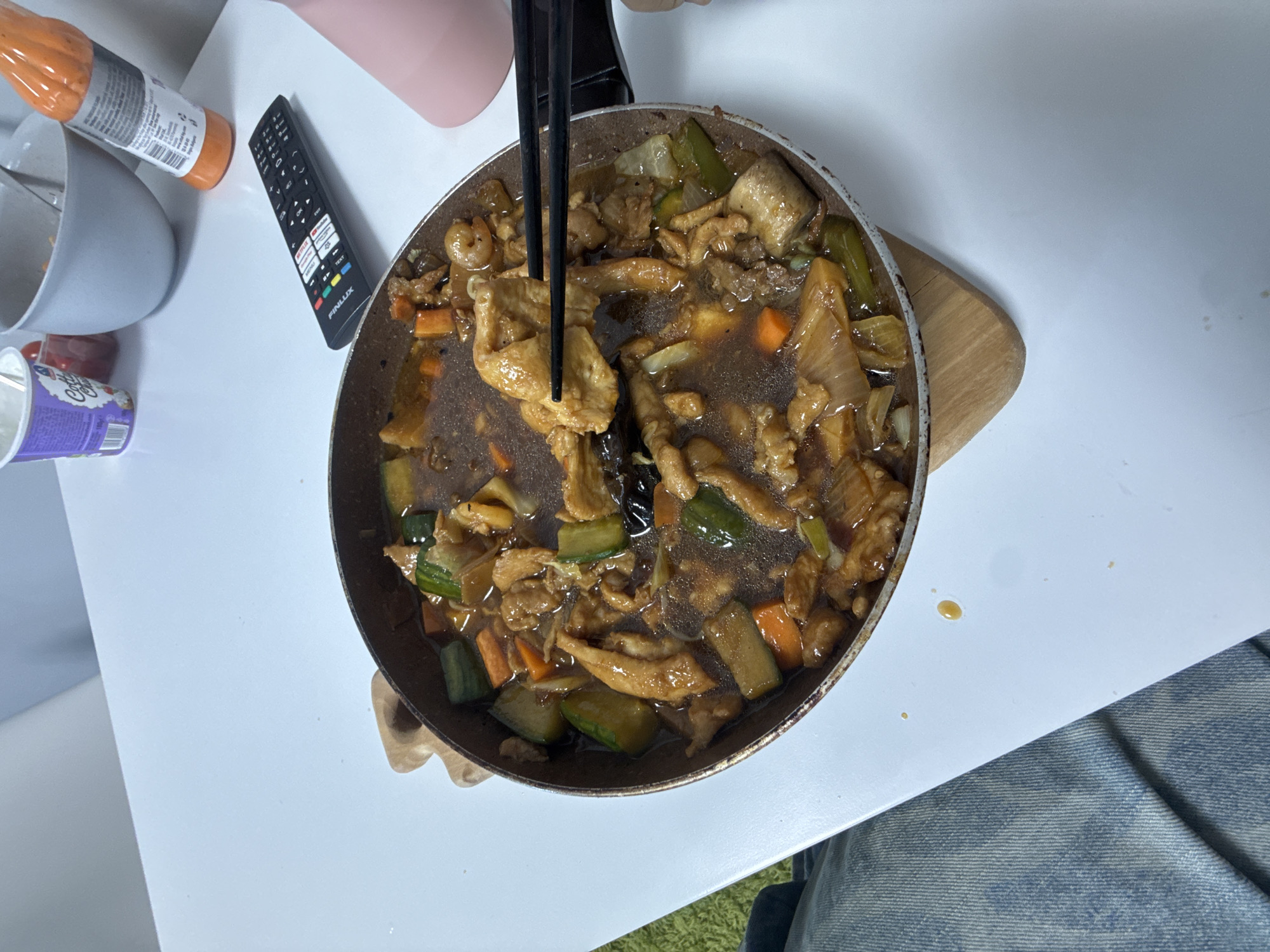 A top-down shot shows a frying pan filled with cooked food, likely a stir-fry, consisting of meat, carrots, and zucchini in a dark sauce. Chopsticks are present, holding up a portion of the food. The pan rests on a wooden cutting board placed on a white surface. To the left are a remote control, a hot sauce bottle, and a yogurt cup. In the lower right corner, a person's lap is visible, wearing denim jeans. The location is likely a domestic setting. There is no indication of Sofia, Bulgaria.
